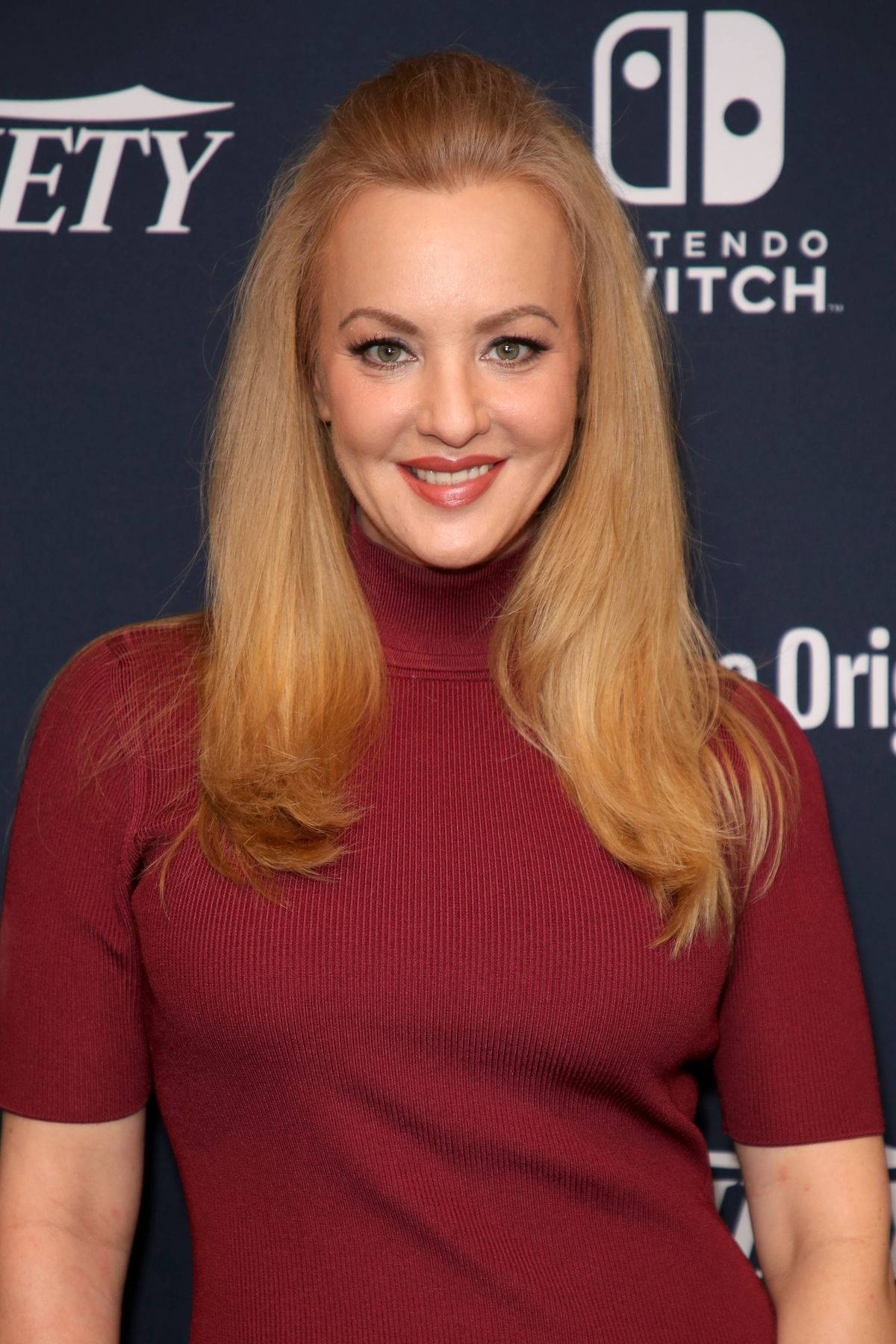 Celebrity 桌布 壁紙 — 一位金髮微笑的女性穿著紅色高領毛衣，背景為深藍色並帶有Nintendo Switch標誌。