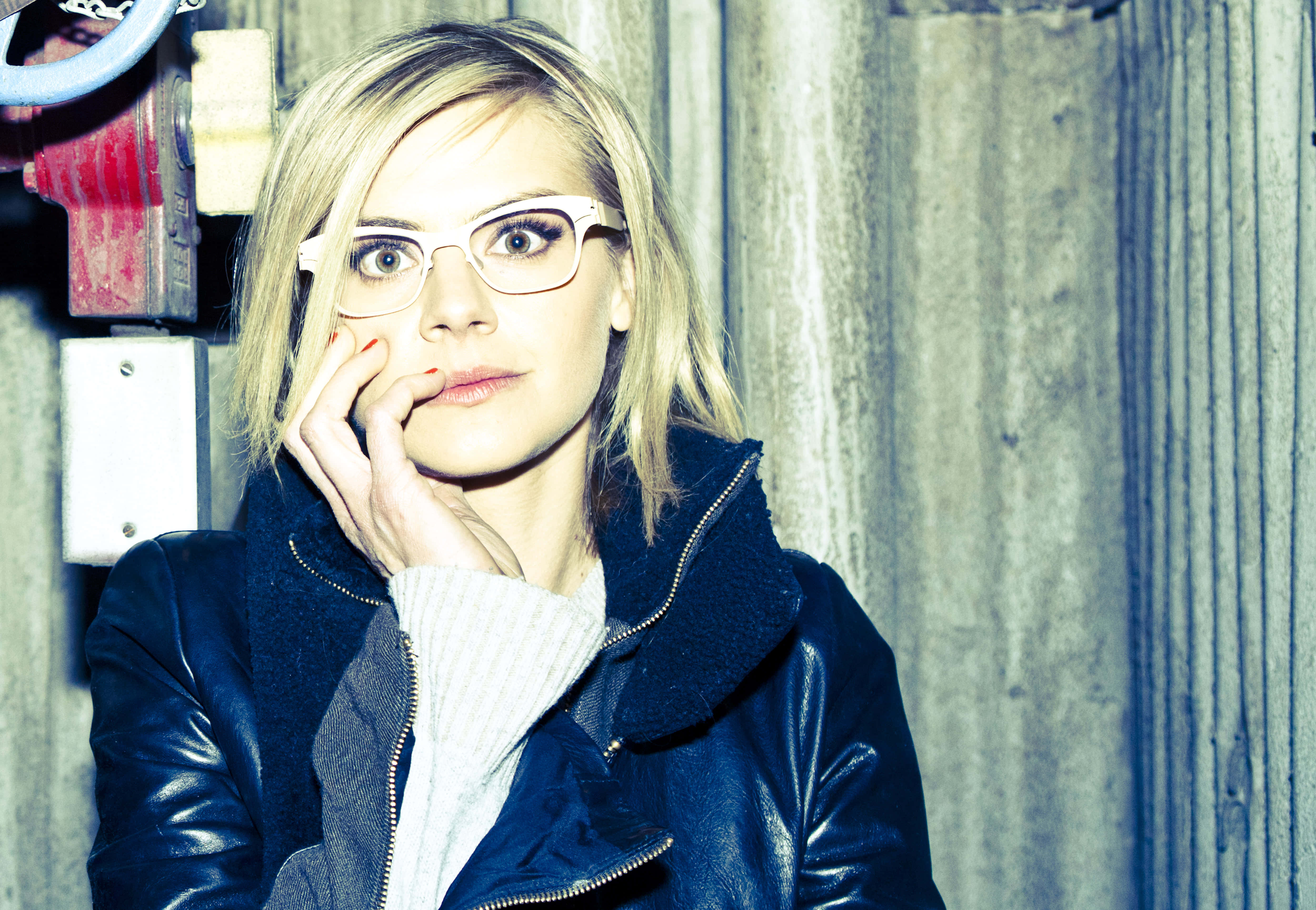 eliza coupe 桌布 壁紙 — 一位金髮戴眼鏡的女性，將下巴擱在手上，靠著風化的木牆，身穿深色皮夾克。