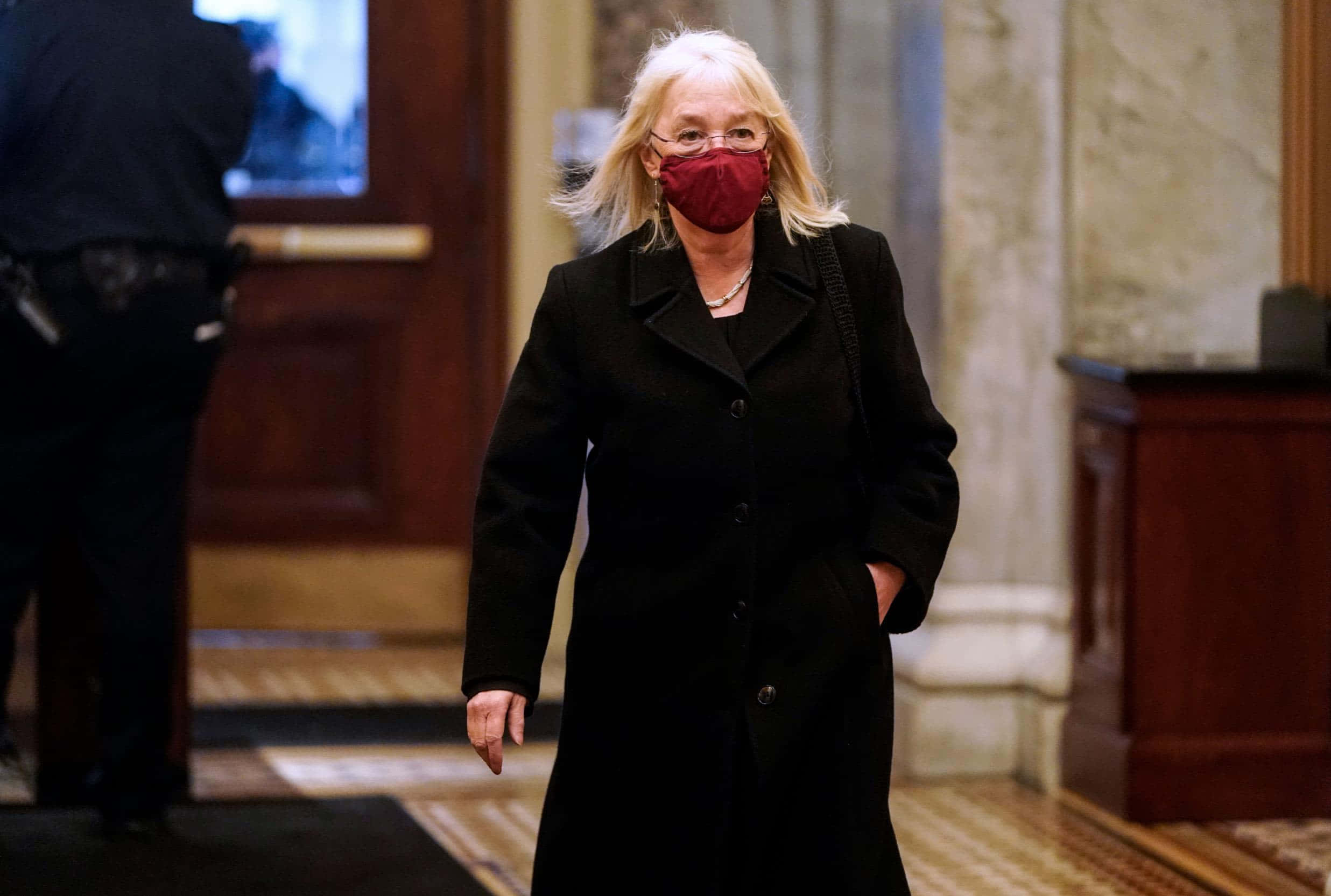 Mask 桌布 壁紙 — 一位金髮戴眼鏡的女性，自信地穿著黑色外套，戴著紅色口罩，走過宏偉的大理石和木質走廊。