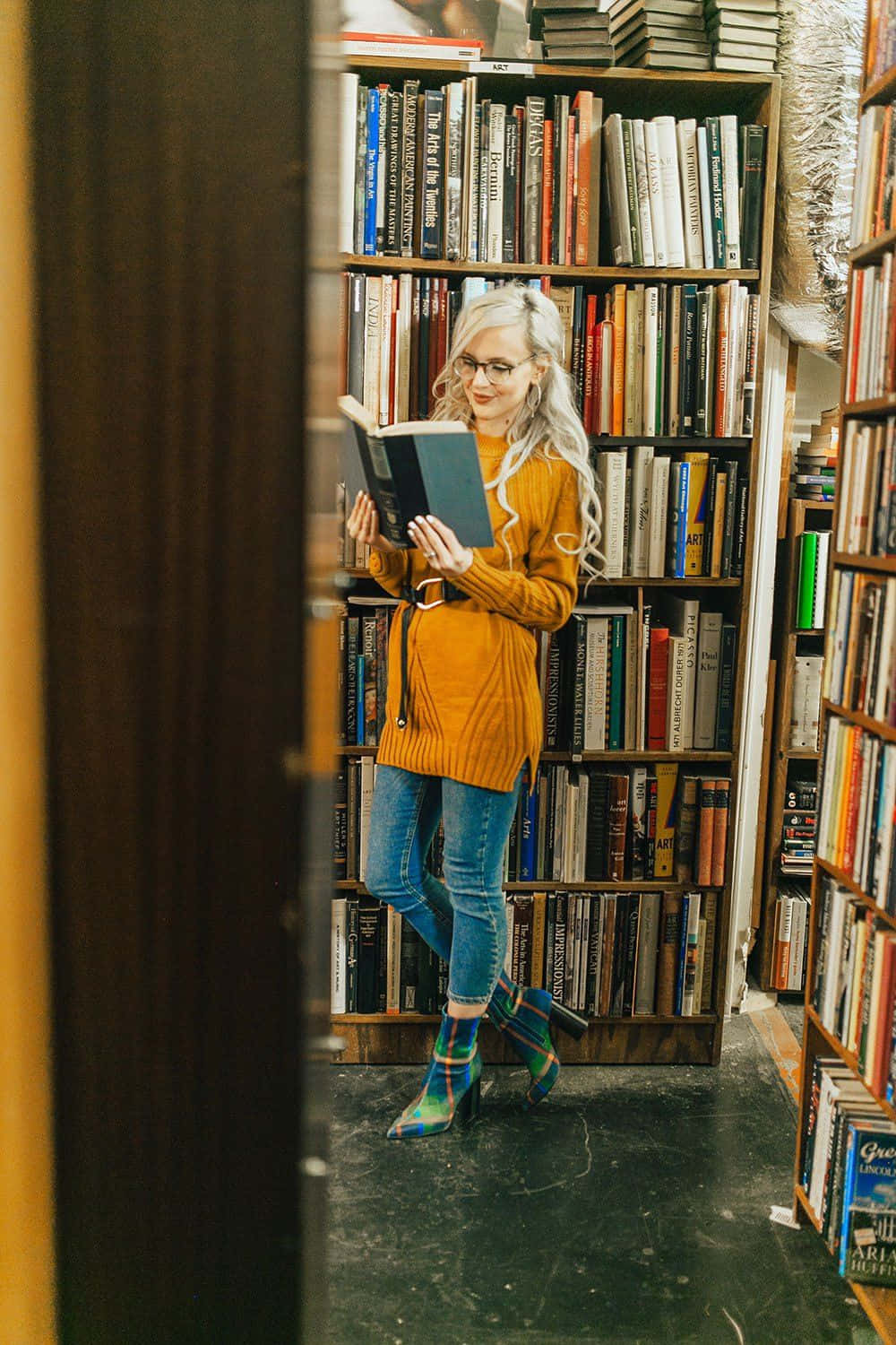 Library 桌布 壁紙 — 一位金髮戴眼鏡的女性在舒適的書店通道中閱讀一本書，通道兩旁是高高的木質書架，擺滿了書籍。
