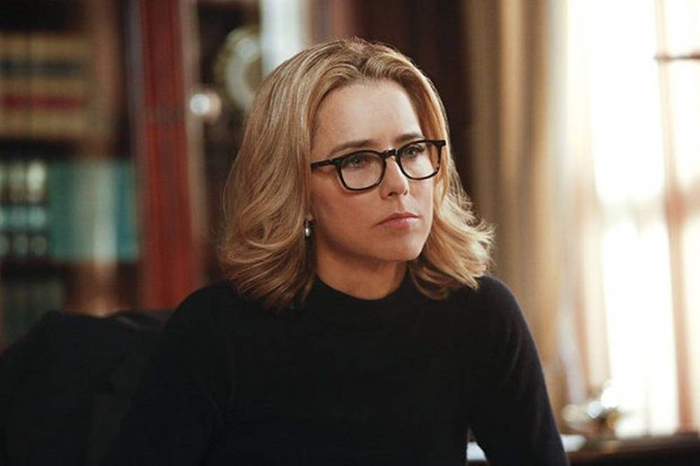 Tea Leoni 桌布 壁紙 — 一位金髮戴眼鏡的女性坐在一個溫暖照明的房間裡，身後有書架和窗簾。