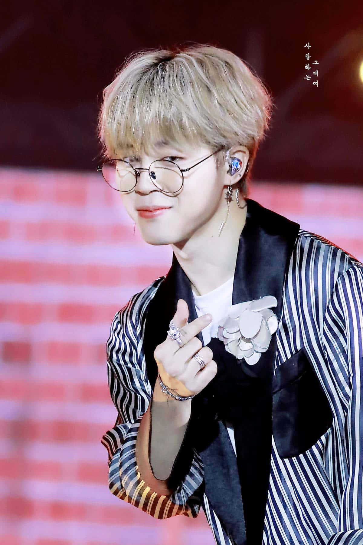 BTS 桌布 壁紙 — 一位金髮戴眼鏡的年輕男子在紅磚舞台上微笑比出和平手勢。