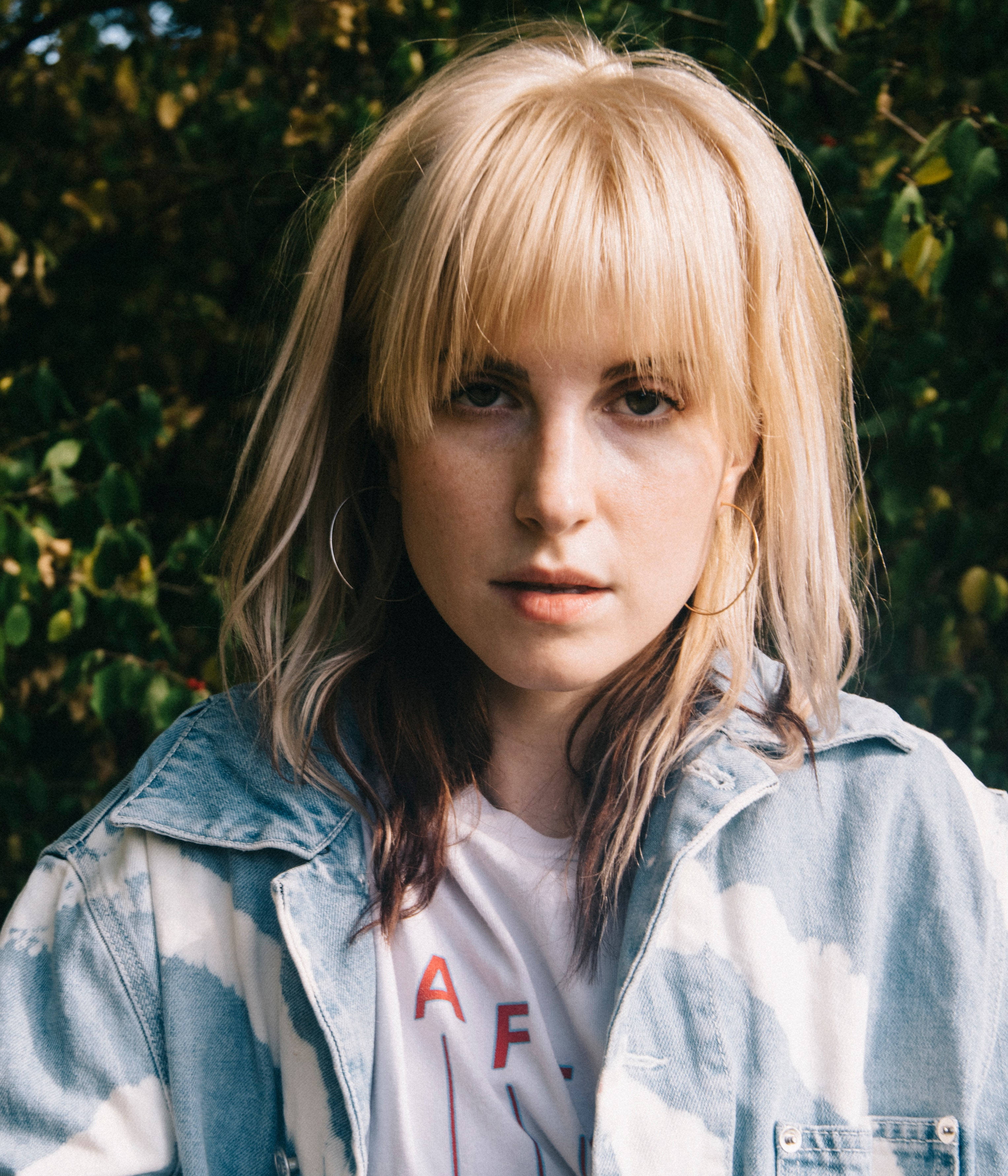 Hayley Williams 桌布 壁紙 — 一位金髮戴耳環的年輕女子直視鏡頭，身穿紮染牛仔外套，背景是茂密的綠色樹葉。