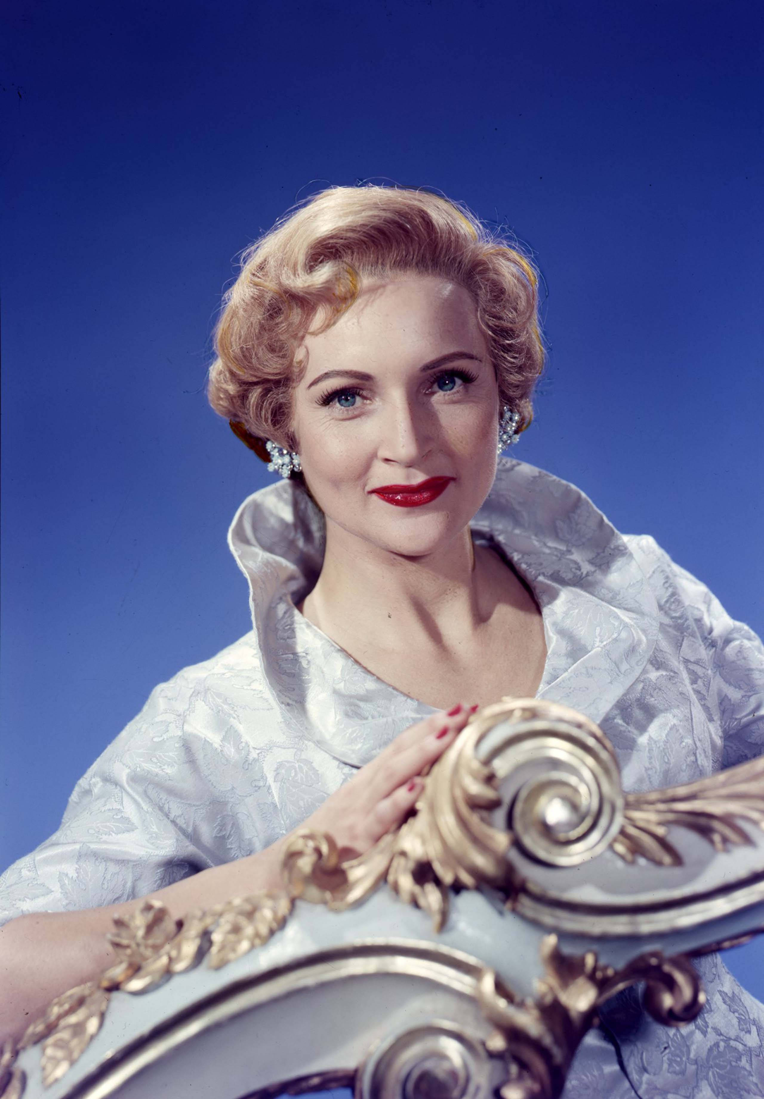 Betty White 桌布 壁紙 — 一位金髮捲曲、紅唇迷人的女士，在深藍色背景下優雅地擺姿勢，手放在華麗的金色椅子上。
