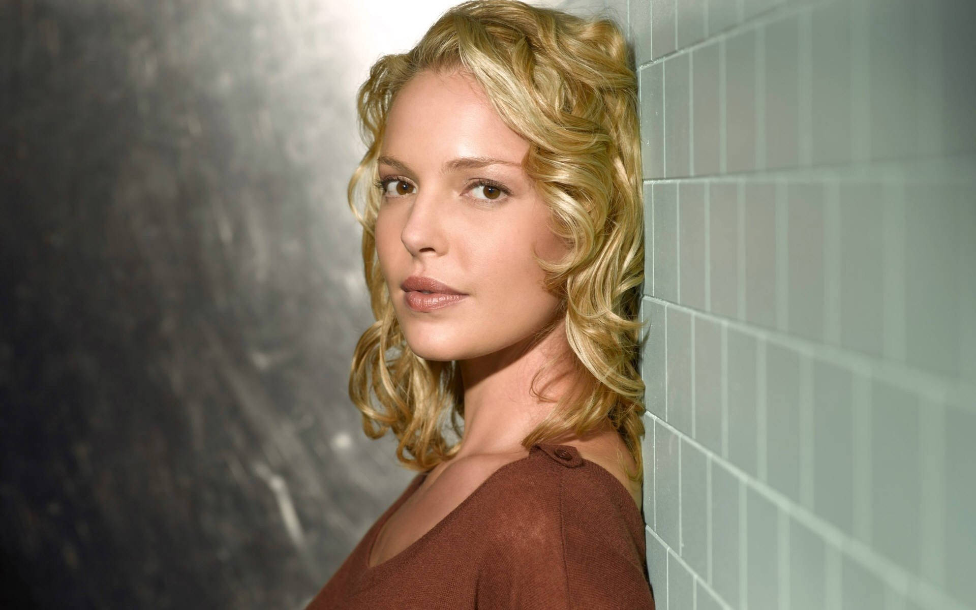 Katherine Heigl 桌布 壁紙 — 一位金髮捲髮女士靠著磁磚牆，在柔和光線下直視鏡頭。