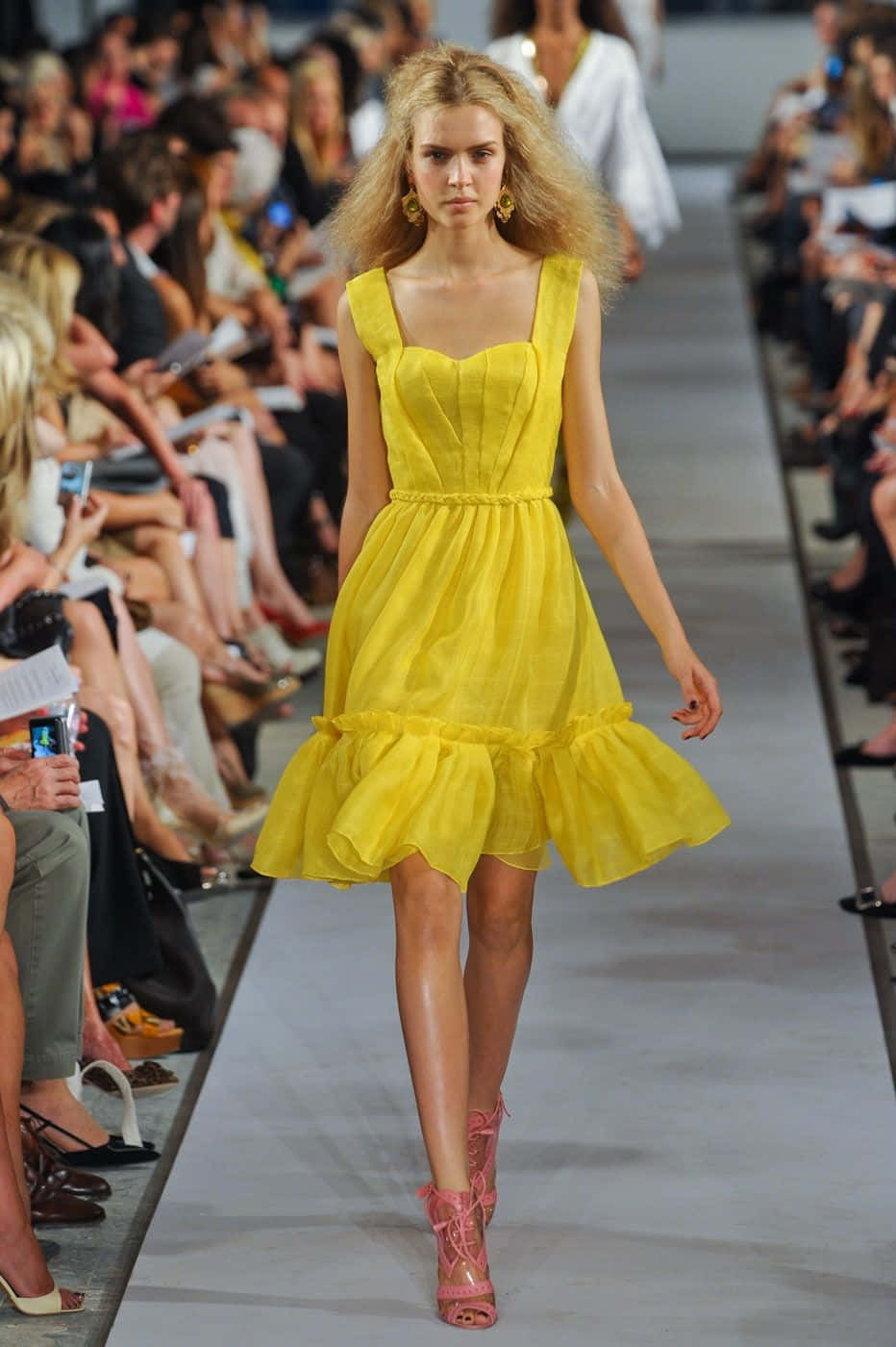 Yellow Fashion 桌布 壁紙 — 一位金髮模特兒穿著鮮黃色荷葉邊連衣裙，在兩旁坐滿觀眾的T台上自信地走著。