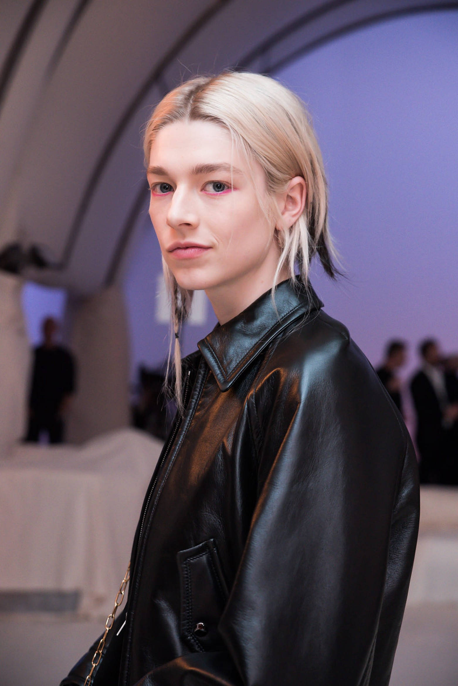 Hunter Schafer 桌布 壁紙 — 一位金髮模特兒穿著黑色皮夾克，在柔和的紫色環境燈光下平靜地凝視鏡頭。
