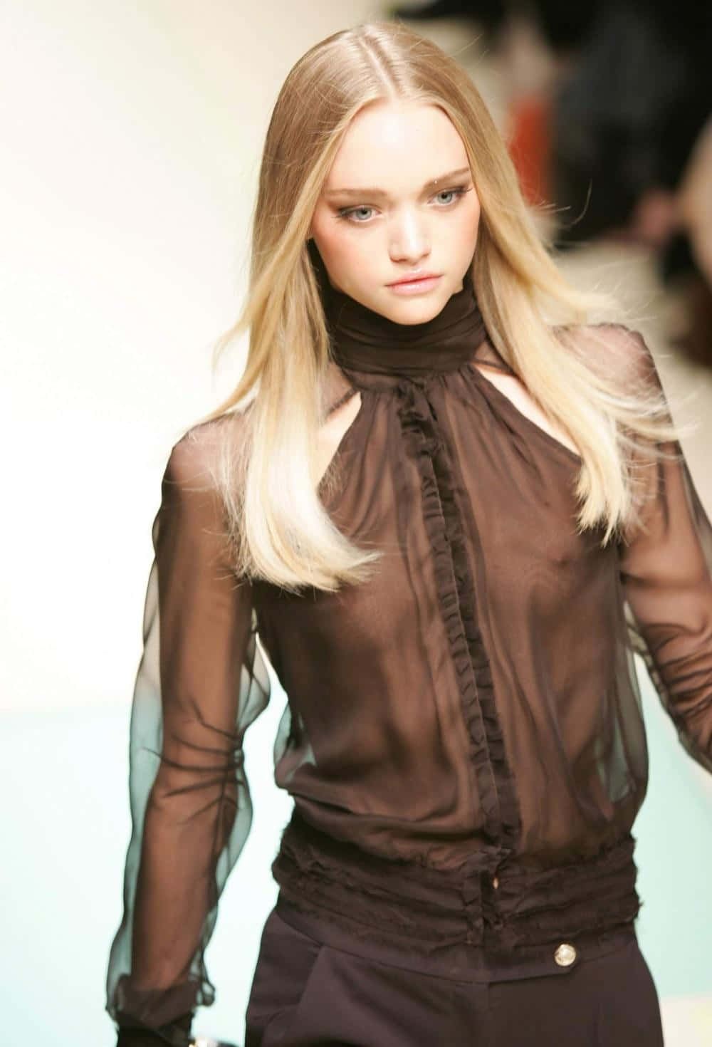 Gemma Ward 桌布 壁紙 — 一位金髮模特兒自信地走在伸展台上，身穿帶荷葉邊細節的透視棕色襯衫和配套褲子。