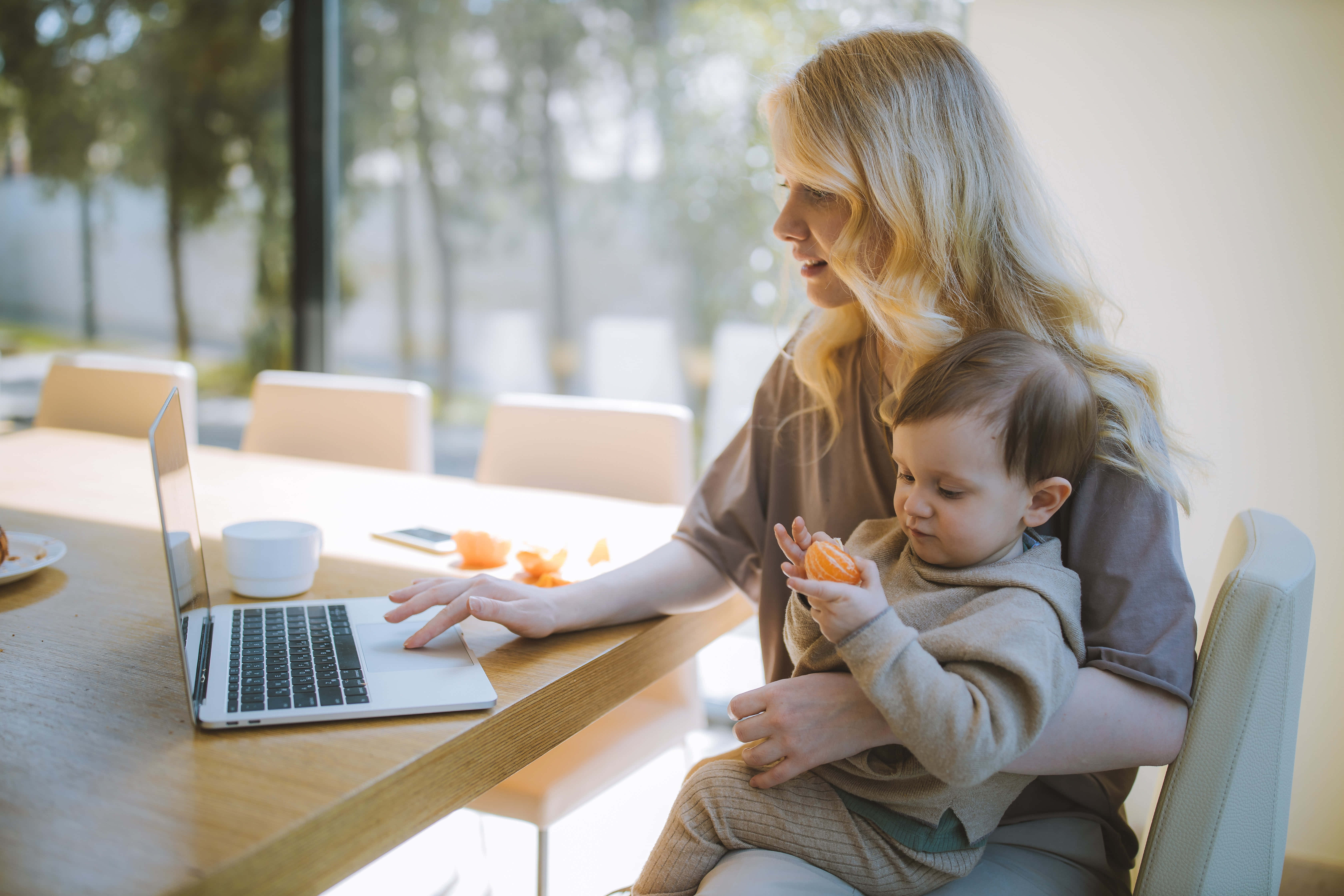 Mom 桌布 壁紙 — 一位金髮母親坐在木桌旁使用筆記型電腦，同時抱著正在吃柳橙片的小孩。