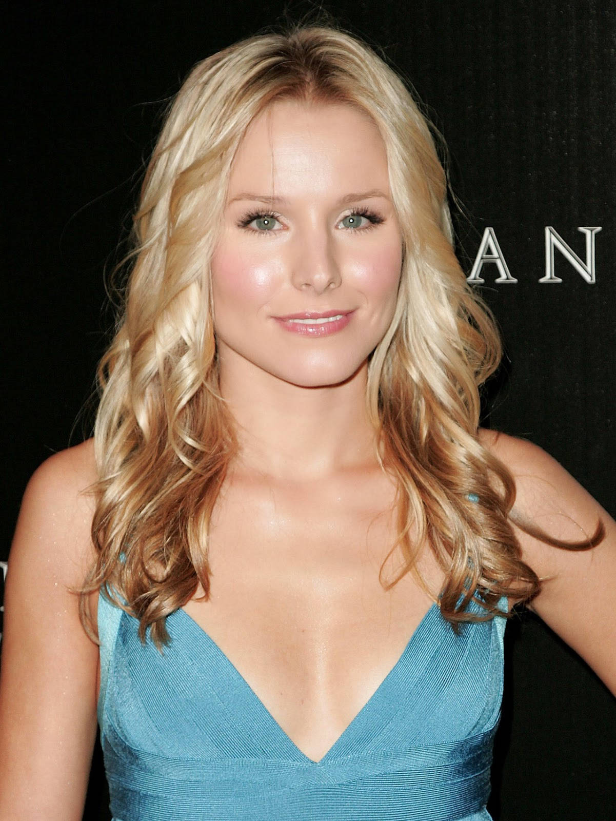 kristen bell 桌布 壁紙 — 一位金髮波浪長髮的女性，身穿藍色連衣裙，在深色背景下溫柔微笑。