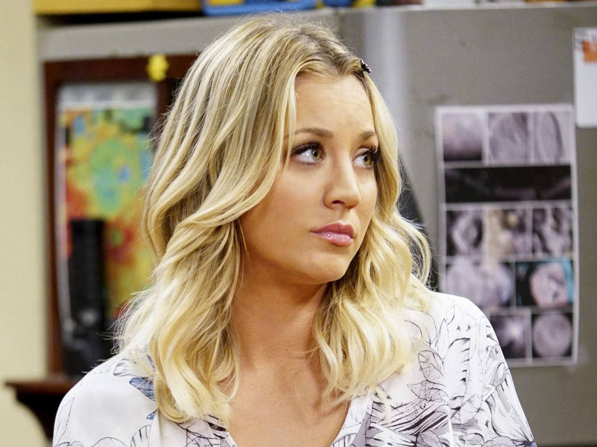 Kaley Cuoco 桌布 壁紙 — 一位金髮波浪長髮的女性在一個有冰箱和牆上照片的房間裡望向鏡頭外。
