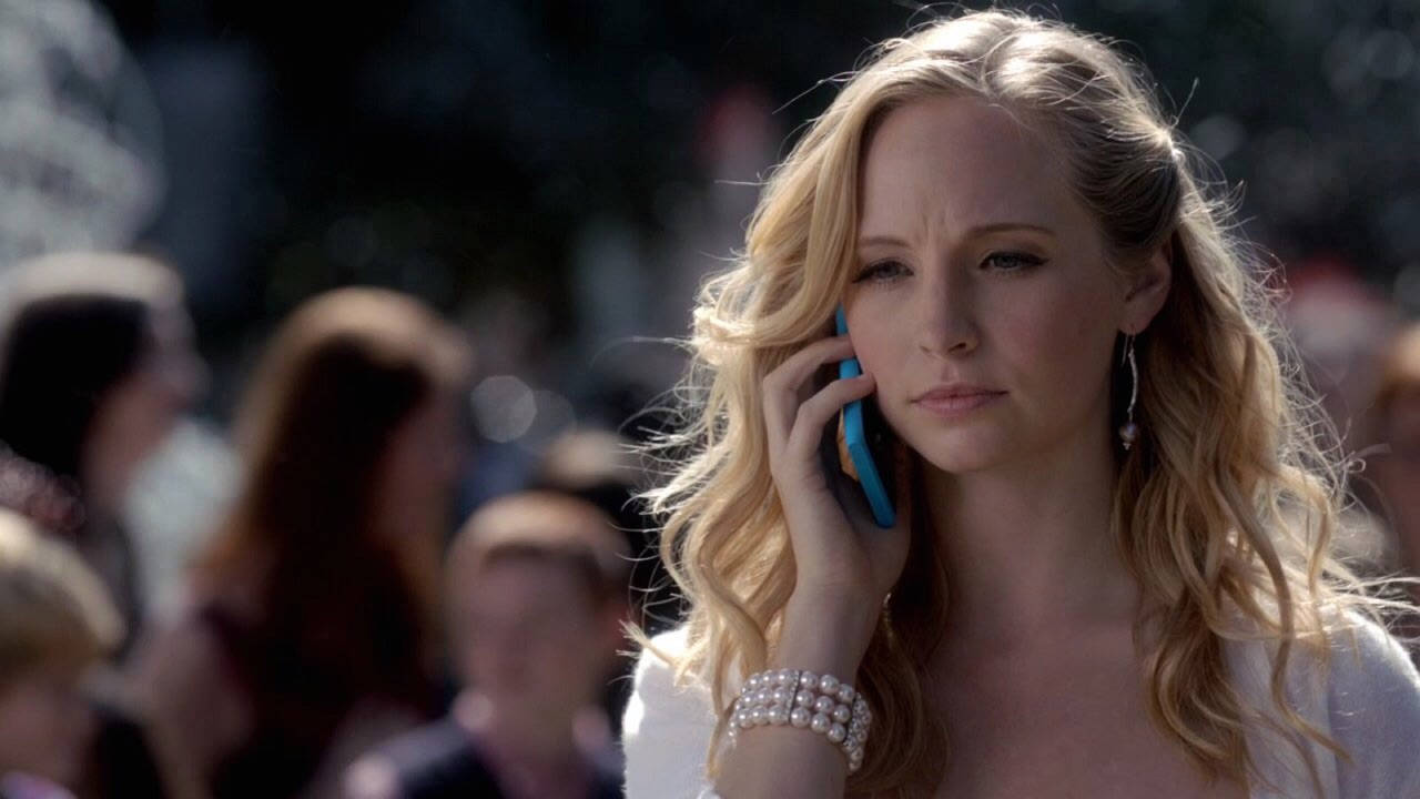 caroline 桌布 壁紙 — 一位金髮波浪長髮的女性在陽光普照的人群中，戴著珍珠手鐲，用藍色電話通話。