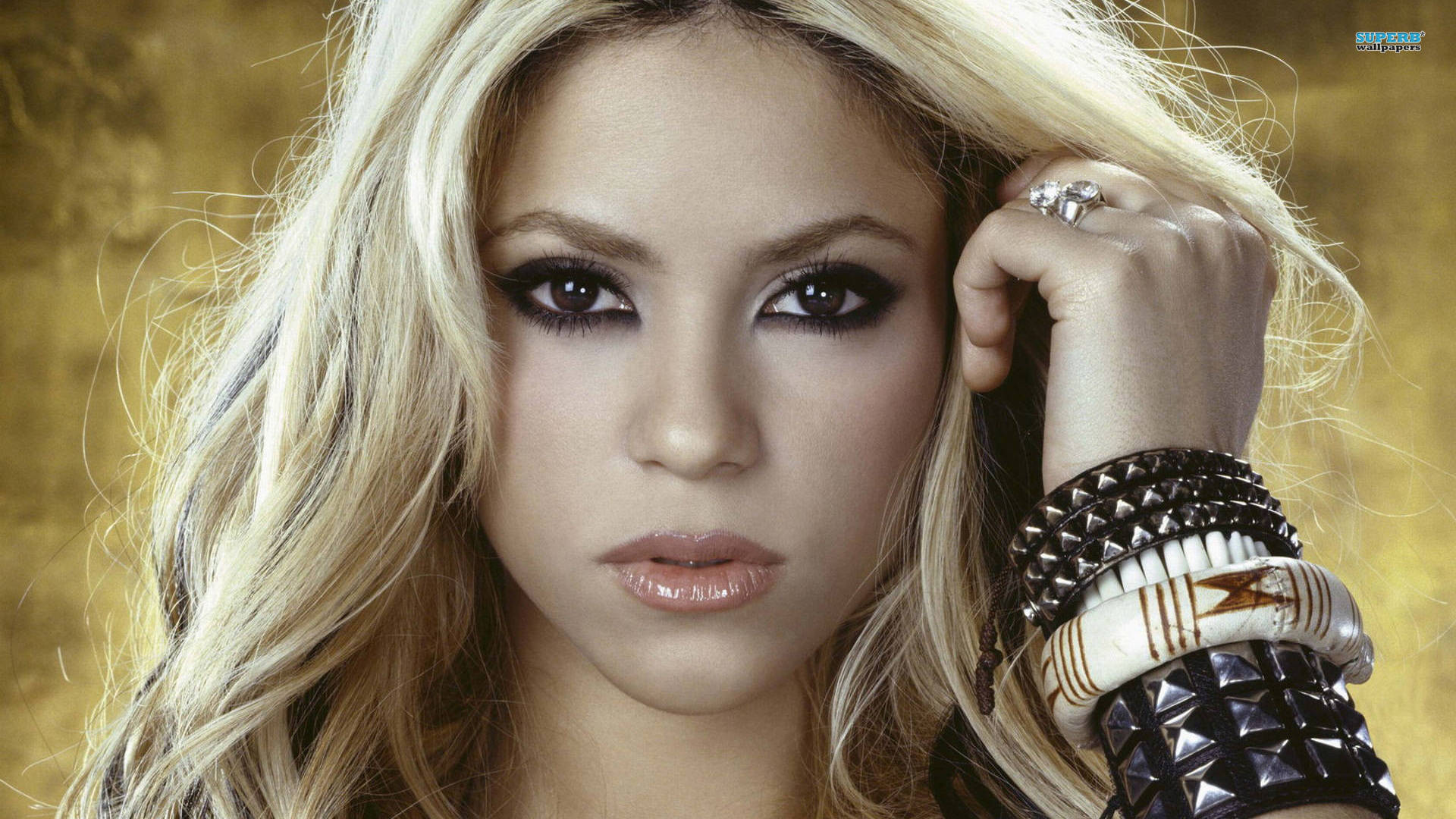 Shakira 桌布 壁紙 — 一位金髮濃妝的迷人女性，在金色紋理背景前佩戴尖刺手鐲擺姿勢。