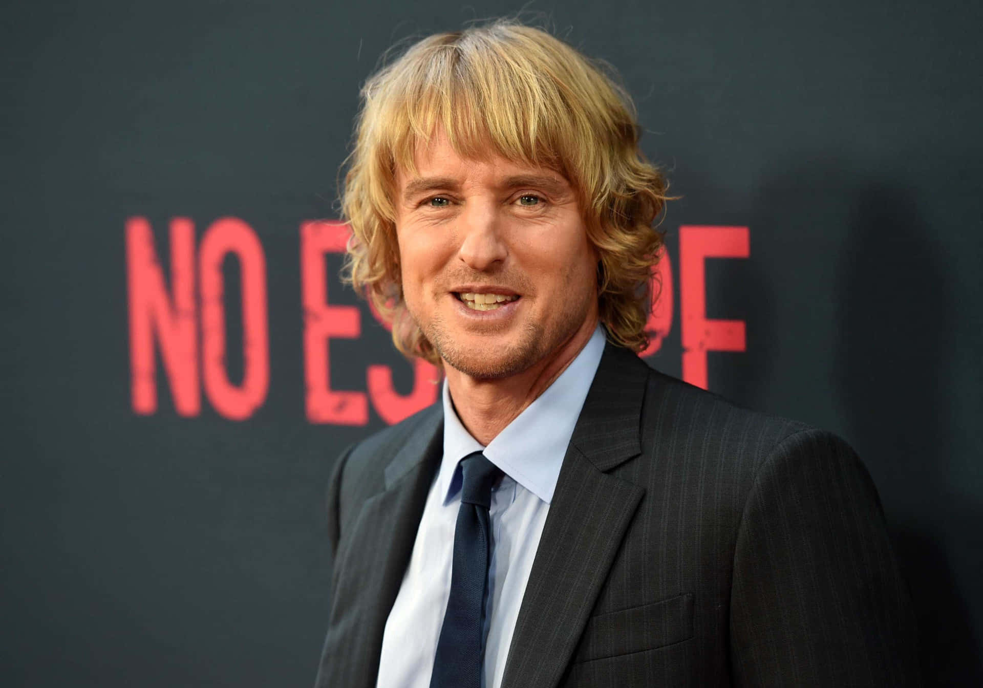 Owen Wilson 桌布 壁紙 — 一位金髮男子穿著深色西裝，在帶有紅色文字的深色背景前對著鏡頭微笑。