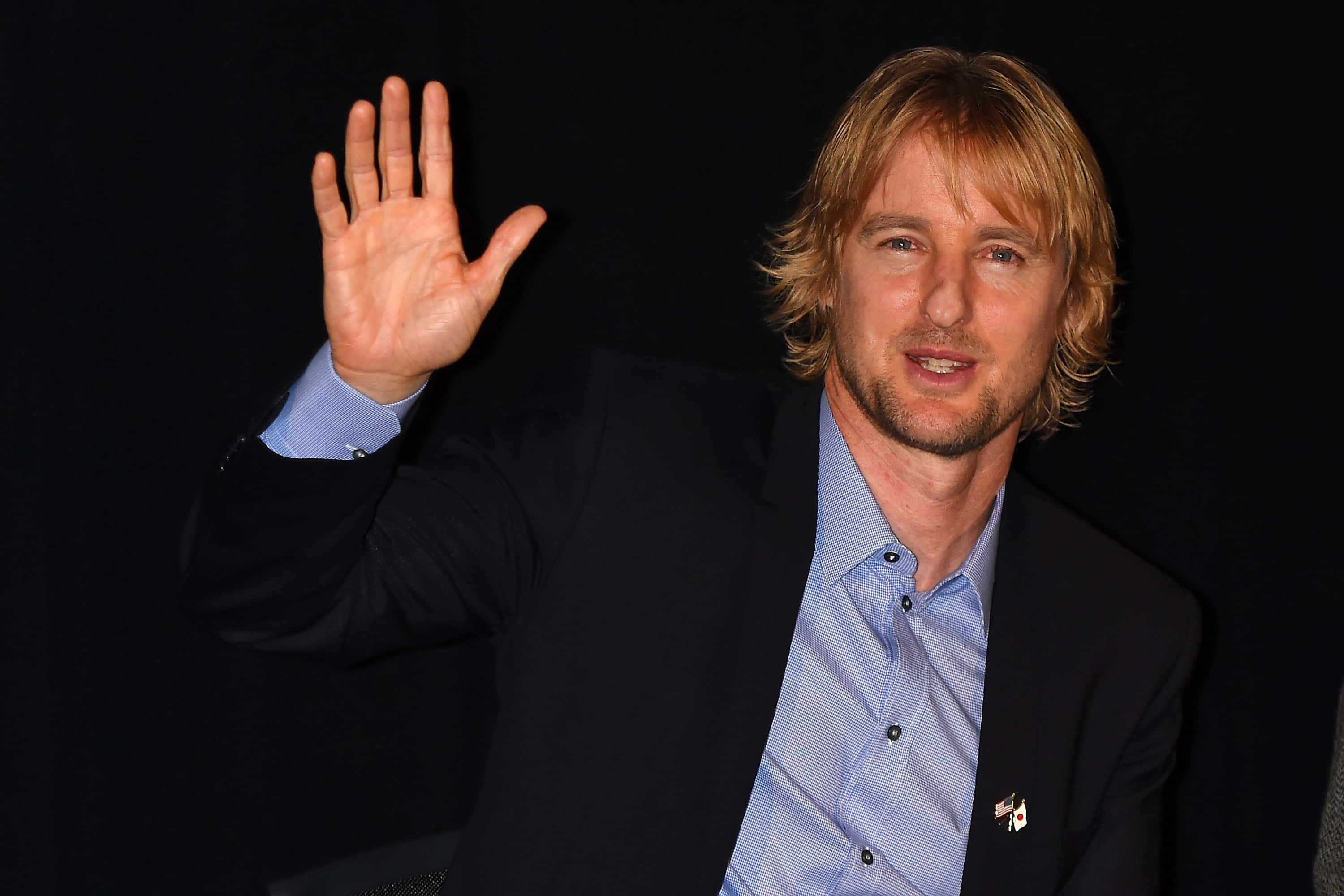 Owen Wilson 桌布 壁紙 — 一位金髮男子穿著深色西裝和藍色襯衫，在黑色背景前向鏡頭揮手。