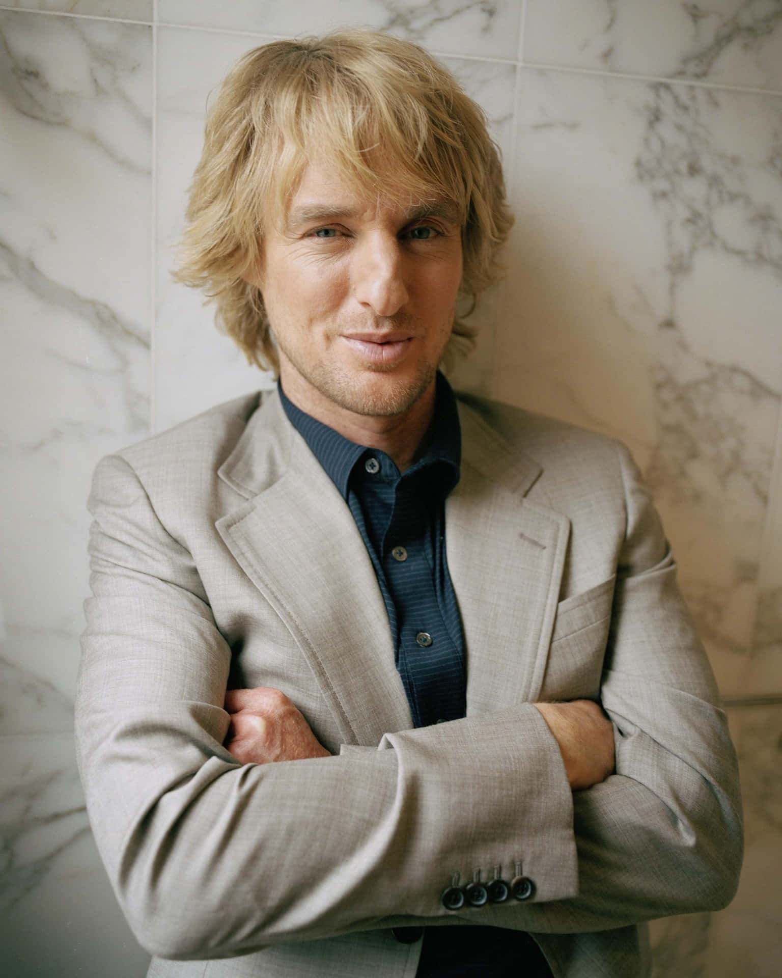 Owen Wilson 桌布 壁紙 — 一位金髮男子身穿米色西裝，雙手交叉放在胸前，自信地靠在大理石牆上。