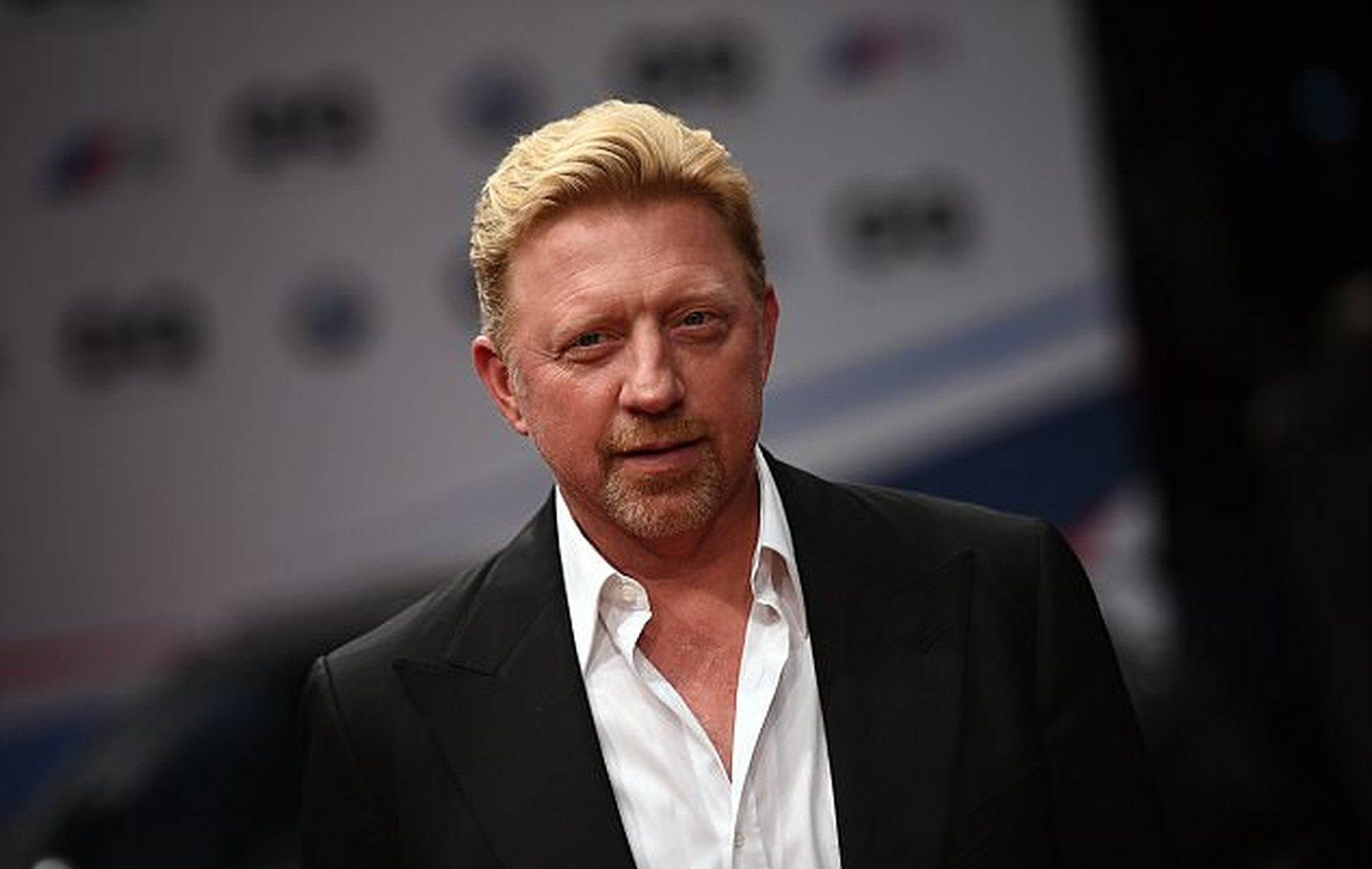 Boris Becker 桌布 壁紙 — 一位金髮留山羊鬍的男人，穿著黑色西裝和白色襯衫，背景模糊帶有品牌標誌。