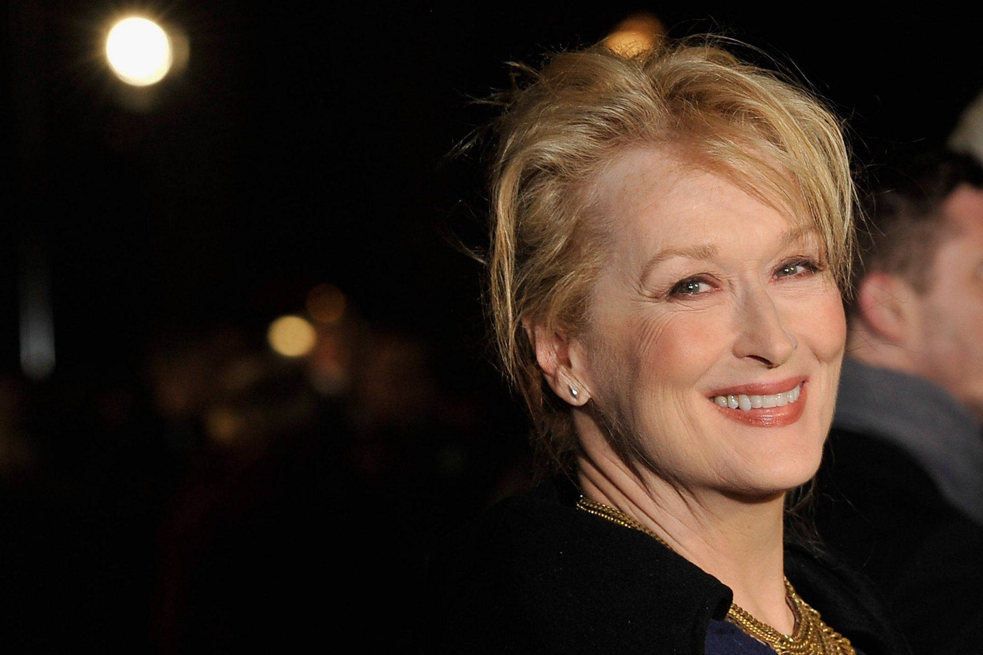 Meryl Streep 桌布 壁紙 — 一位金髮的燦爛女性在黑暗模糊的背景中，於明亮的活動燈光下溫暖地微笑。