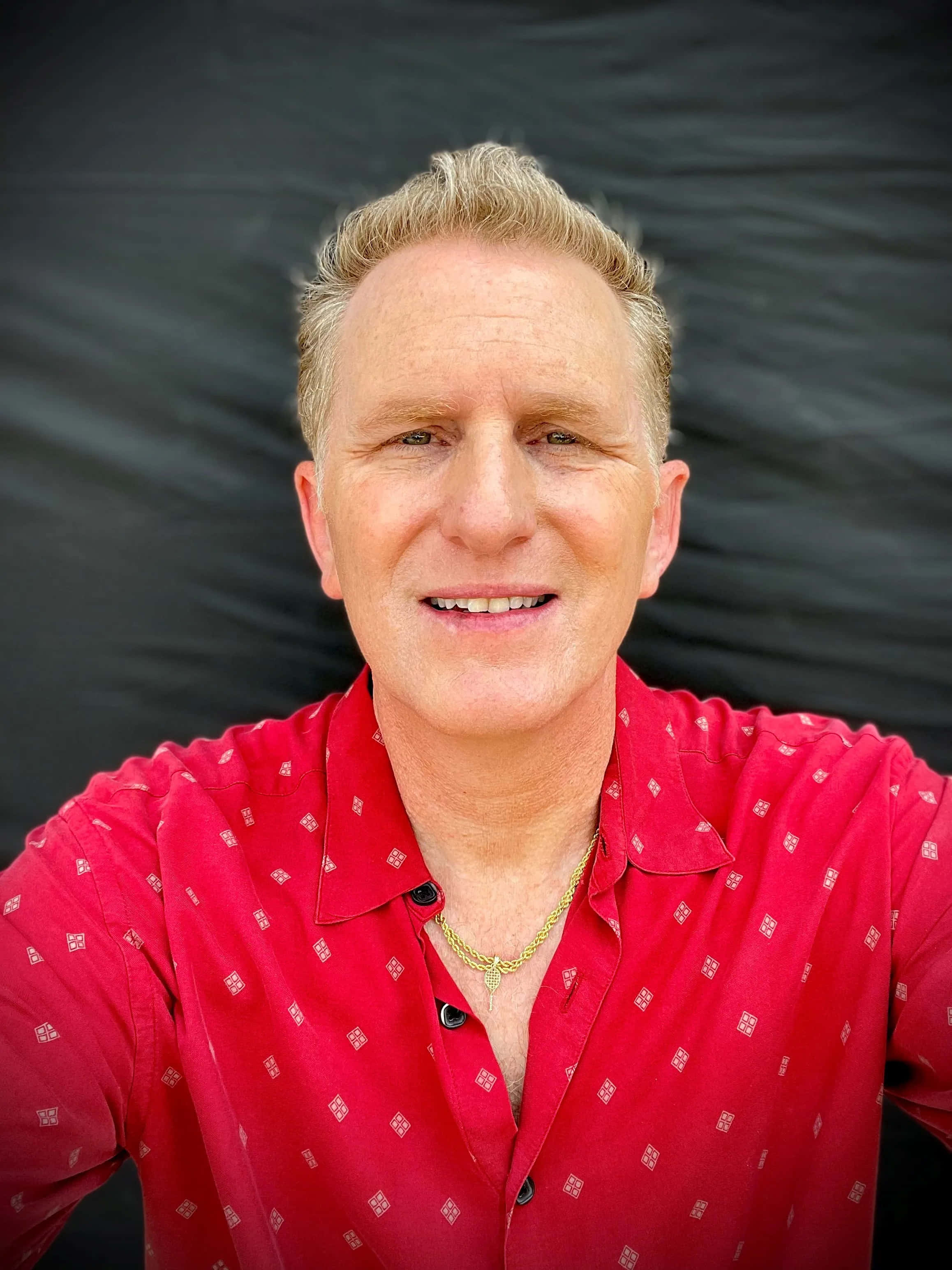 Michael Rapaport 桌布 壁紙 — 一位金髮的開朗男子在深色背景下穿著紅色圖案襯衫溫暖地微笑。