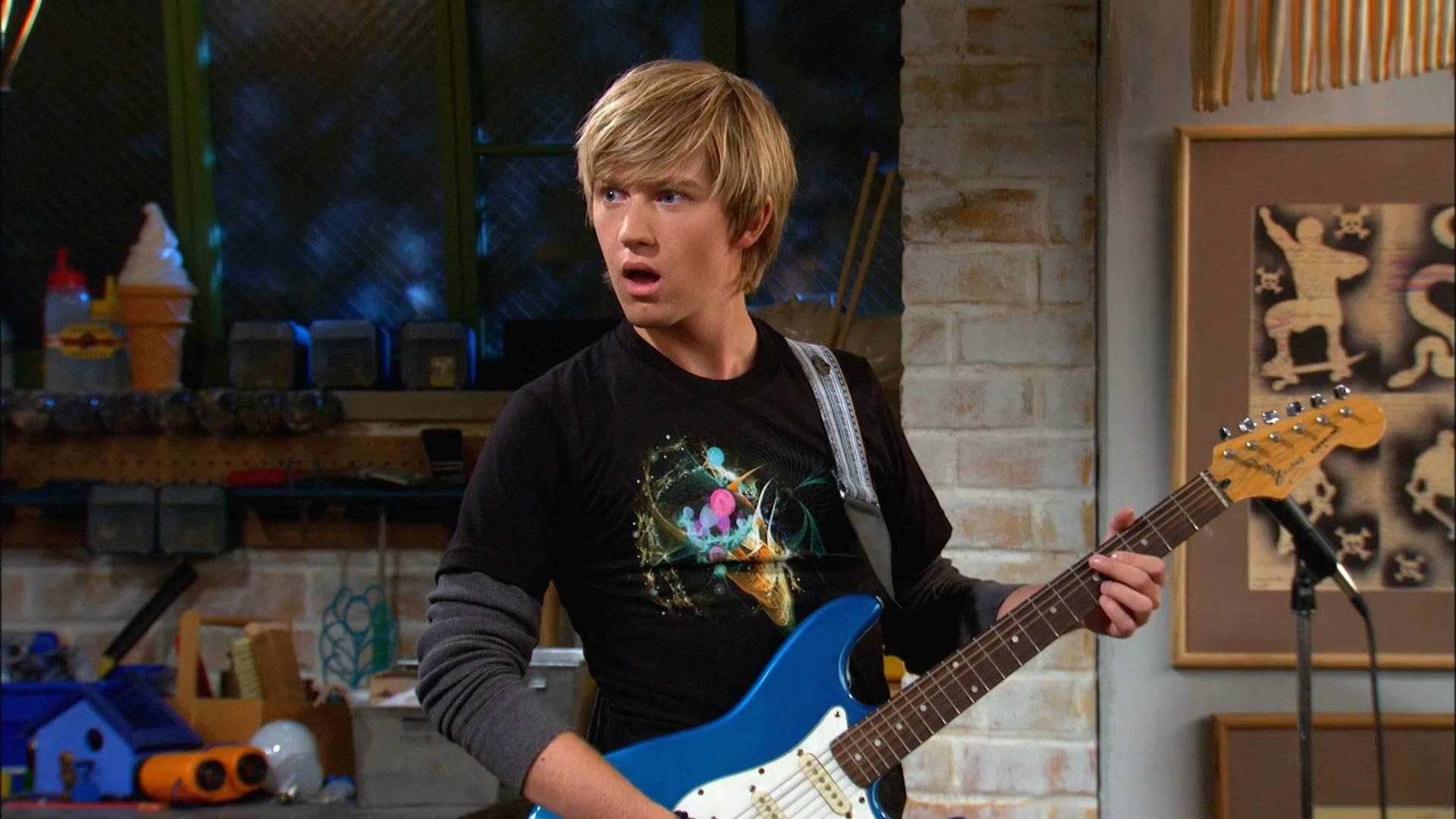 Good Luck Charlie 桌布 壁紙 — 一位金髮的驚訝年輕男子在一個雜亂的車庫工作室裡，拿著一把藍色電吉他，背景有磚牆和裱框藝術品。