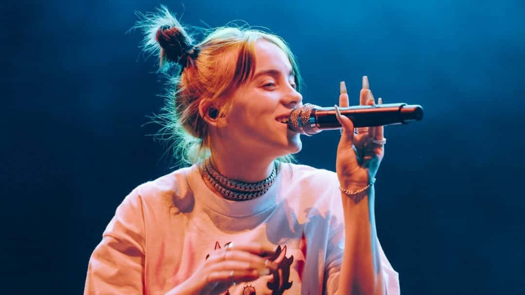 Billie Eilish Laptop 桌布 壁紙 — 一位金髮盤髻的年輕女子在藍色舞台燈光下深情地對著麥克風唱歌。