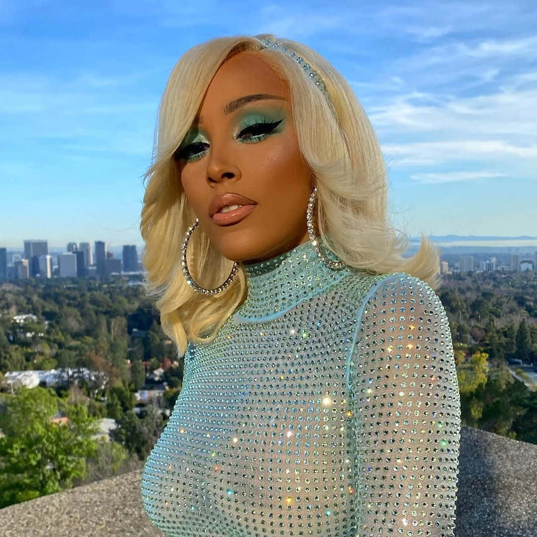 Doja Cat 桌布 壁紙 — 一位金髮碧眼、化著青綠色妝容的迷人女性，穿著閃亮的淺藍色服裝，以城市天際線為背景。