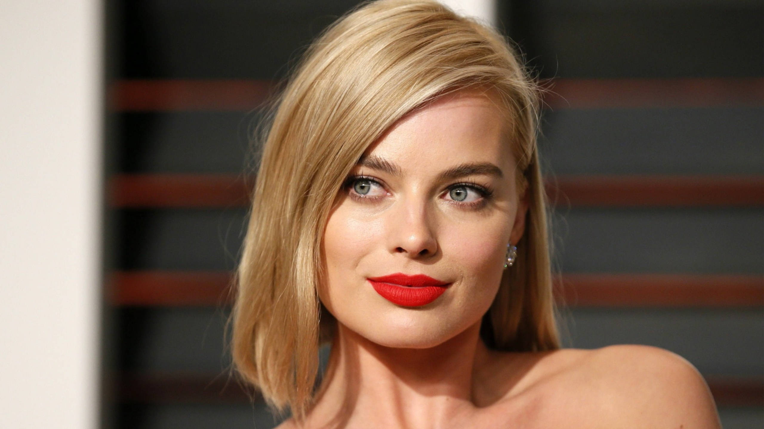 Margot Robbie 桌布 壁紙 — 一位金髮碧眼、塗著大膽紅唇的女性，在模糊的條紋背景前微笑的特寫肖像。