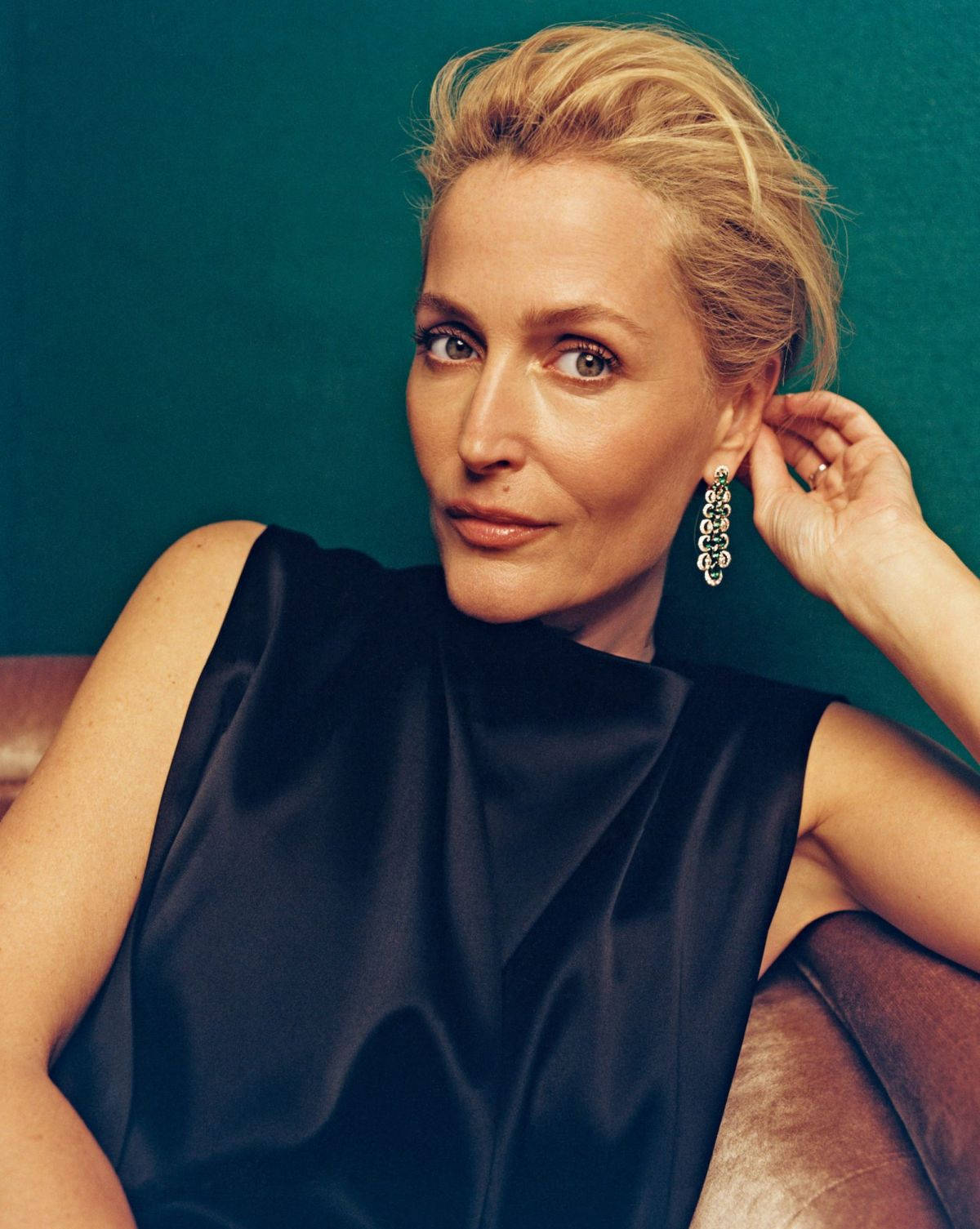 gillian anderson 桌布 壁紙 — 一位金髮碧眼、戴著搶眼耳環的優雅女士，在藍綠色背景前優雅地坐著，呈現奢華的攝影棚肖像。