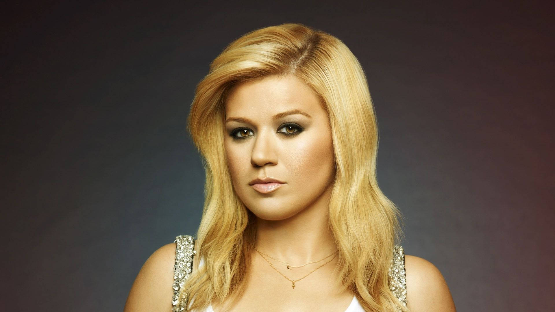 Kelly Clarkson 桌布 壁紙 — 一位金髮碧眼、煙燻妝、穿著閃亮上衣的女性在深色漸變背景前擺姿勢。