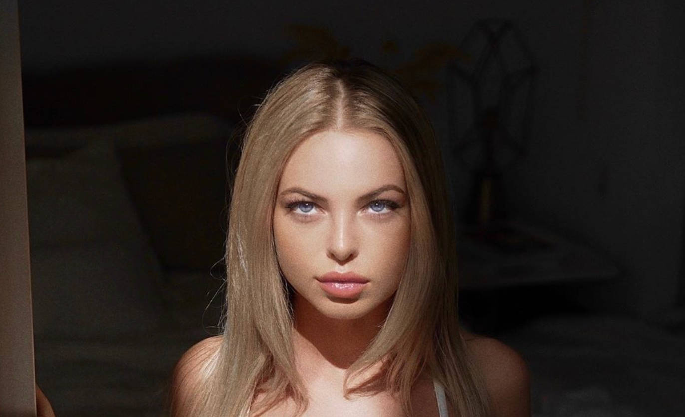 Emma Kotos 桌布 壁紙 — 一位金髮碧眼、目光銳利的女性，在光線昏暗、帶有柔和陰影的房間中直視鏡頭。