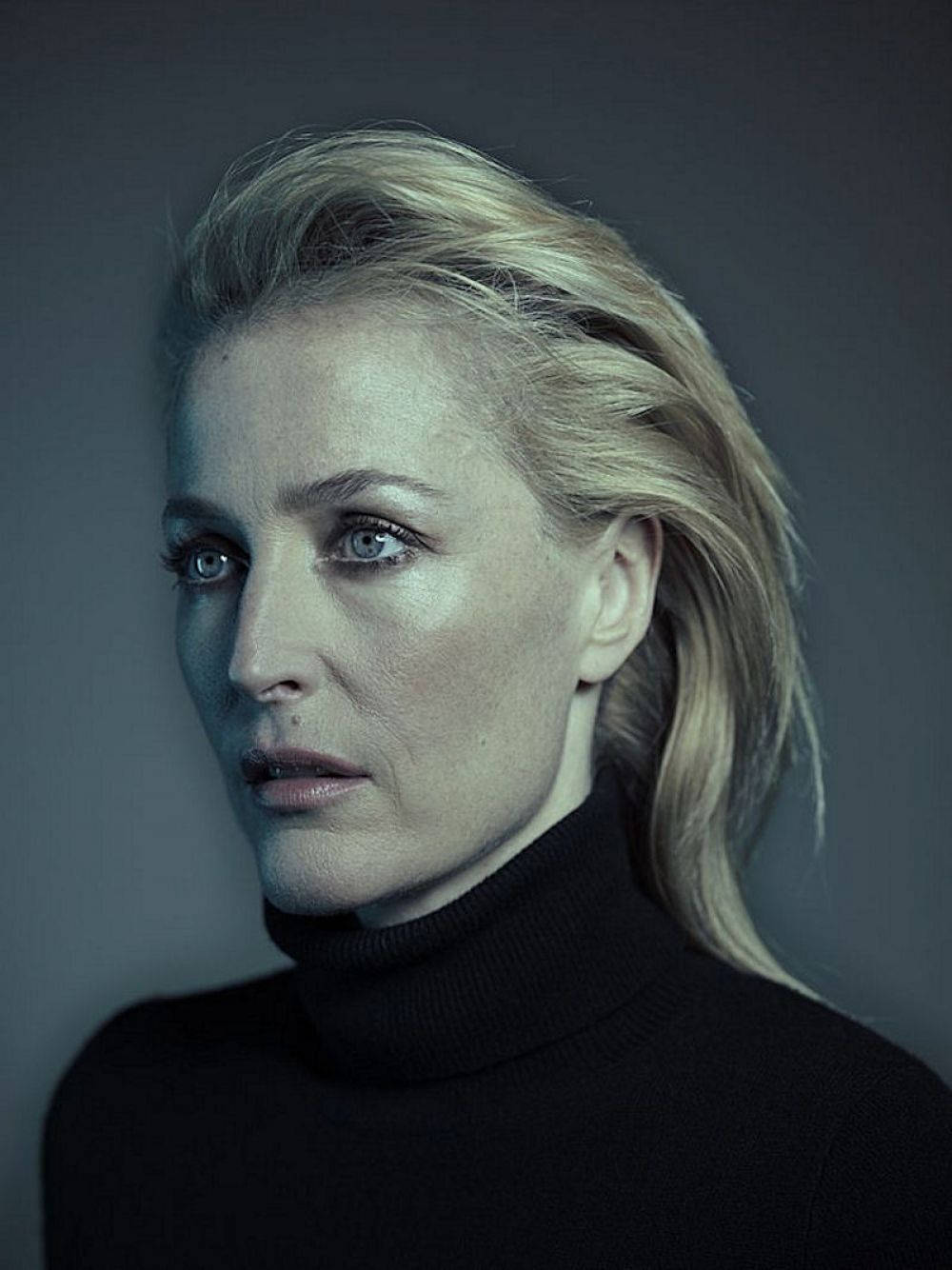 gillian anderson 桌布 壁紙 — 一位金髮碧眼、眼神銳利的女性，在灰色背景前拍攝的陰鬱工作室肖像中專注地凝視。