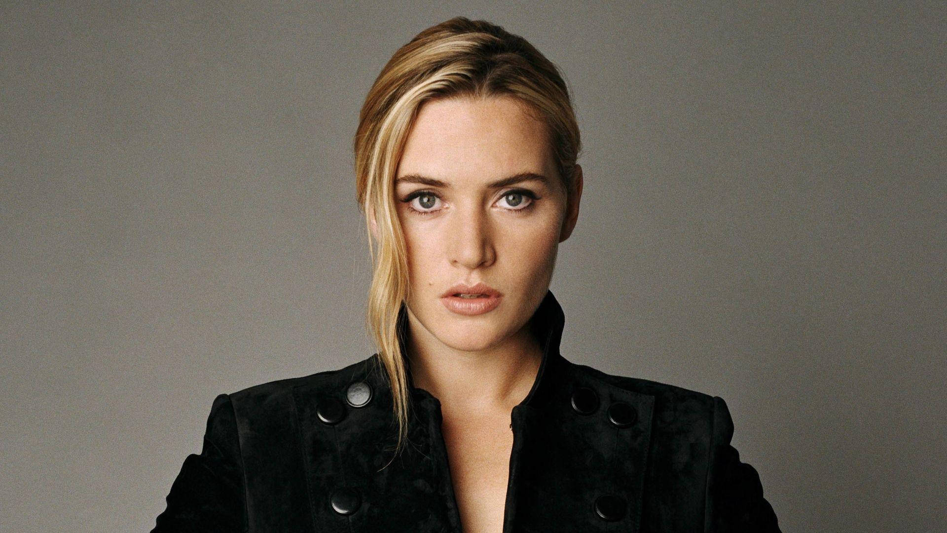 Kate Winslet 桌布 壁紙 — 一位金髮碧眼、眼神銳利的女性，身穿黑色天鵝絨外套，在純灰色背景下直視鏡頭。