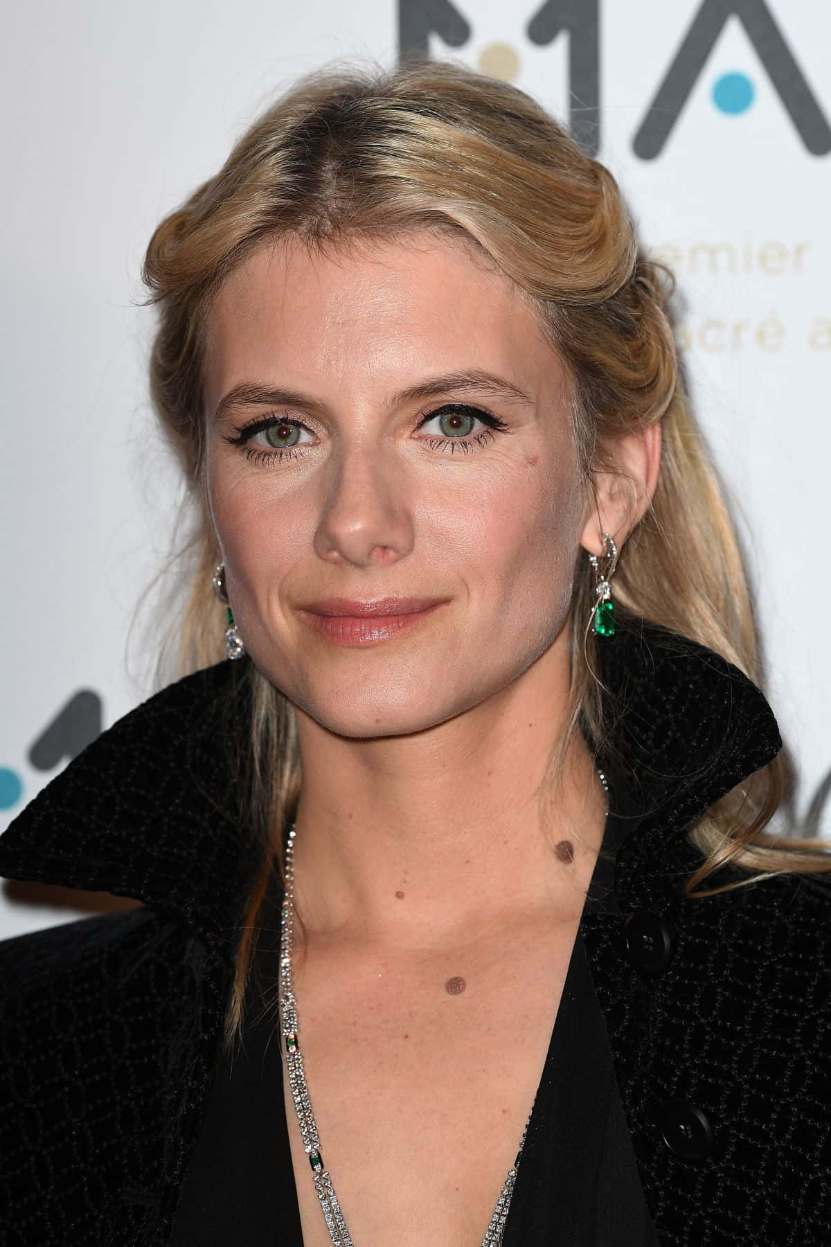 Mélanie Laurent 桌布 壁紙 — 一位金髮碧眼的女人穿著黑色外套和綠色耳環出席活動的特寫肖像。