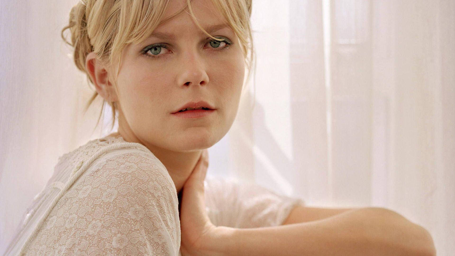 Kirsten Dunst 桌布 壁紙 — 一位金髮碧眼的女性，在柔軟的白色窗簾背景下，用手托著下巴，若有所思地凝視著。