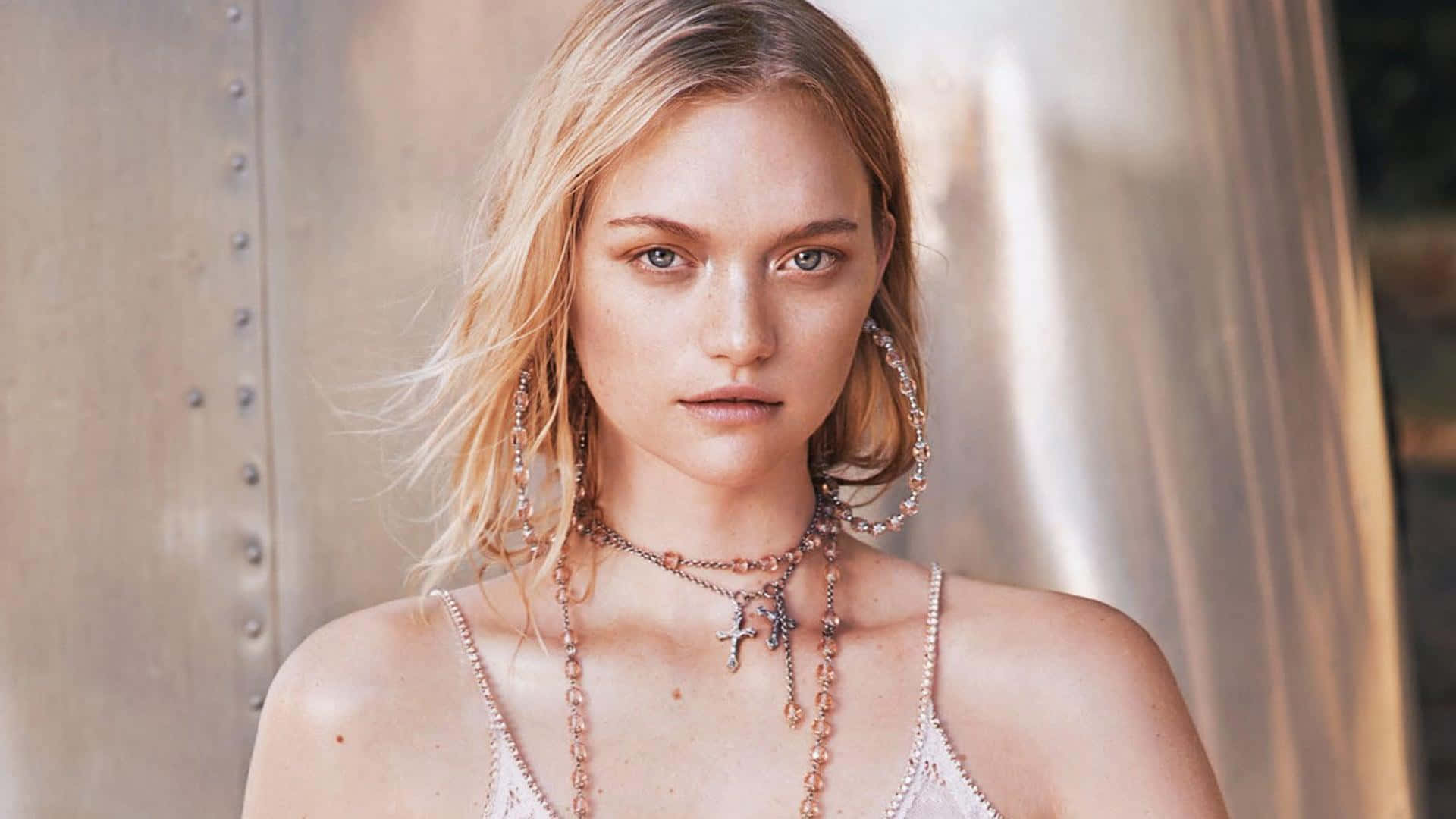 Gemma Ward 桌布 壁紙 — 一位金髮碧眼的女性，在金屬鉚釘背景前佩戴層疊的珍珠和十字架項鍊。
