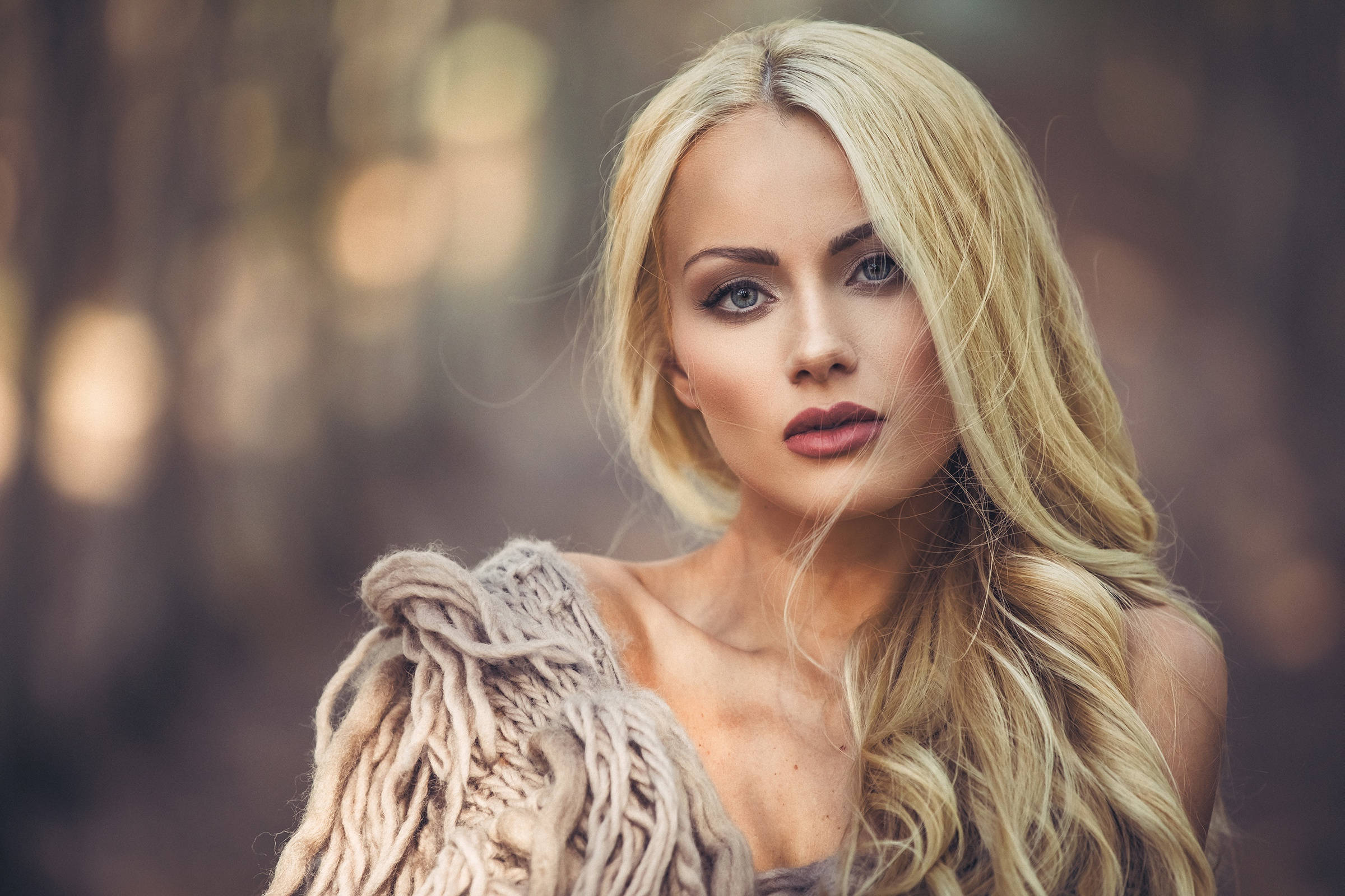 Blonde Woman 桌布 壁紙 — 一位金髮碧眼的女性，披著質感針織披肩，在柔和模糊的森林背景下直視鏡頭。