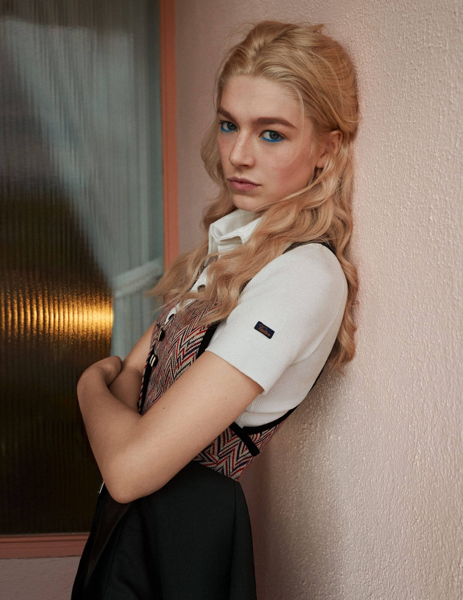 Hunter Schafer 桌布 壁紙 — 一位金髮碧眼的女性，身穿白色襯衫和圖案背心，靠著紋理牆壁。