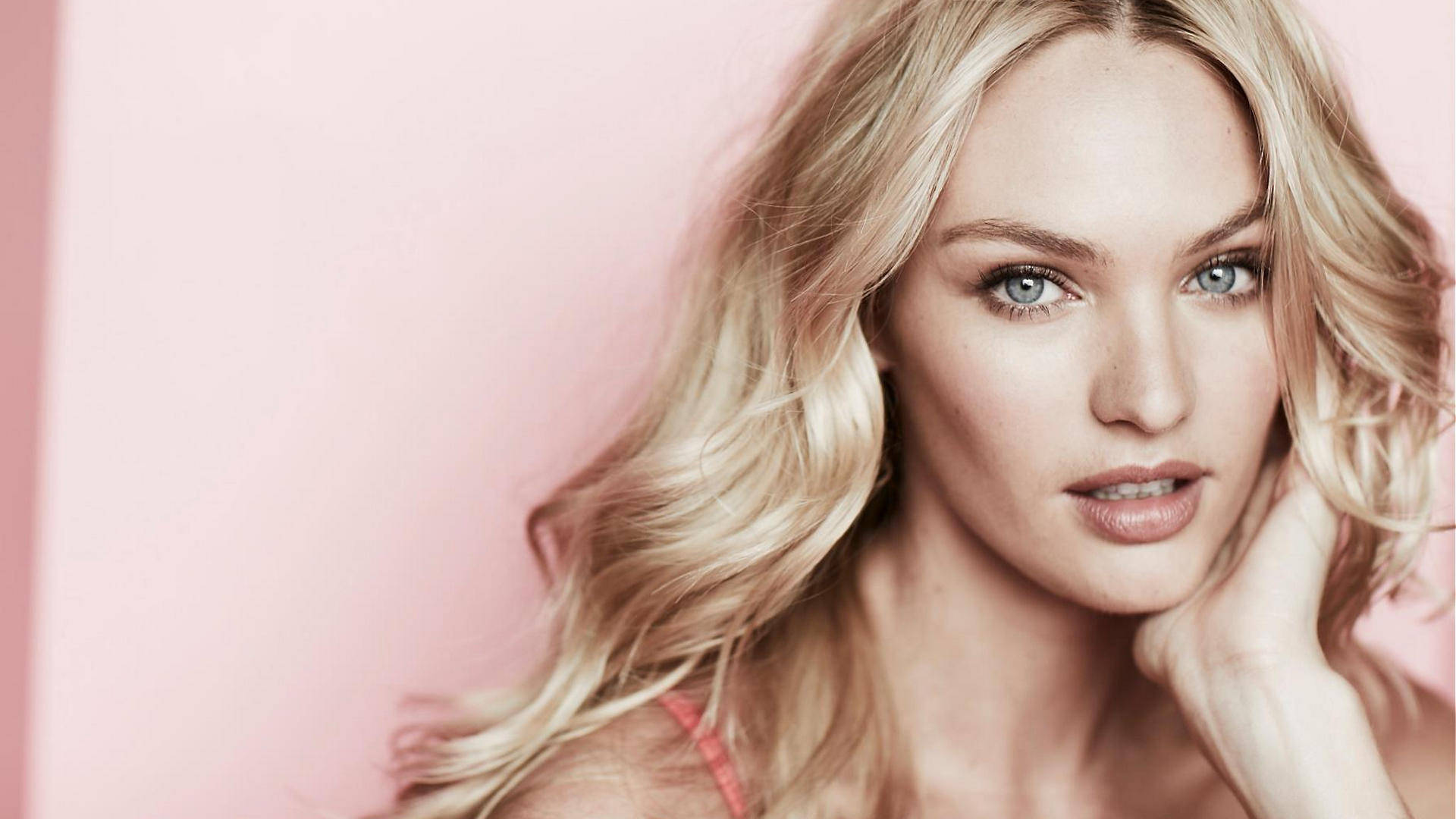 Candice 桌布 壁紙 — 一位金髮碧眼的女性在柔和的粉紅色背景前溫柔地凝視，她的手輕輕地放在臉頰旁。