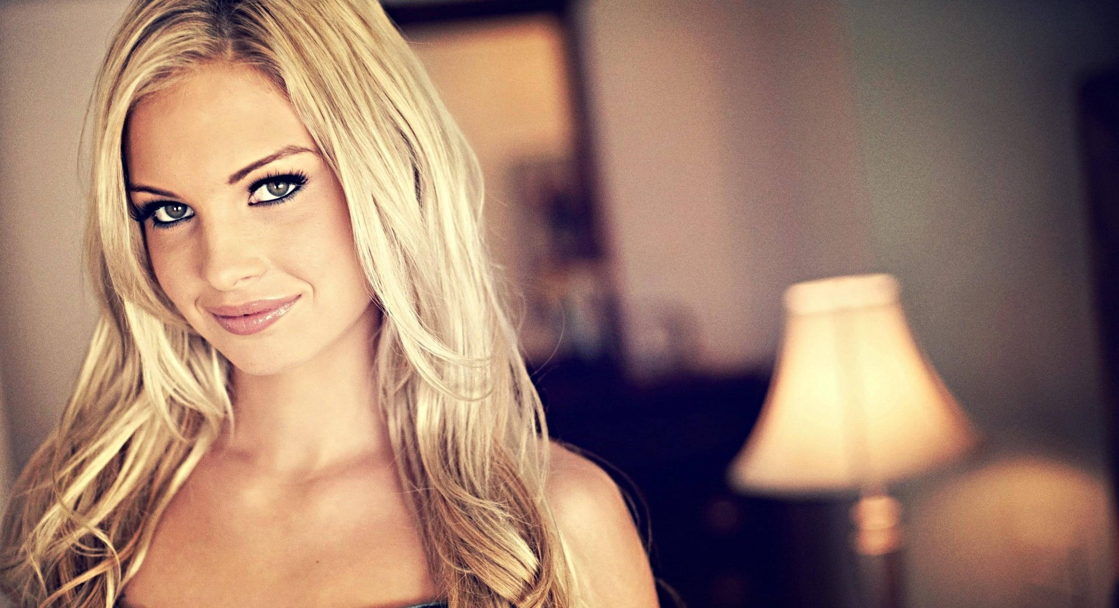 電影 桌布 壁紙 — 一位金髮碧眼的女性在溫暖的室內環境中微笑，背景有一盞發光的燈。
