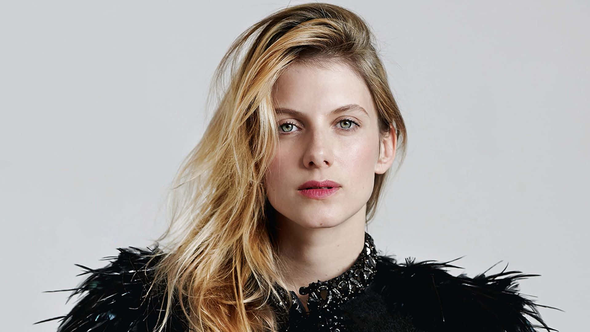 Mélanie Laurent 桌布 壁紙 — 一位金髮碧眼的女性穿著黑色羽毛上衣，背景為純灰色。