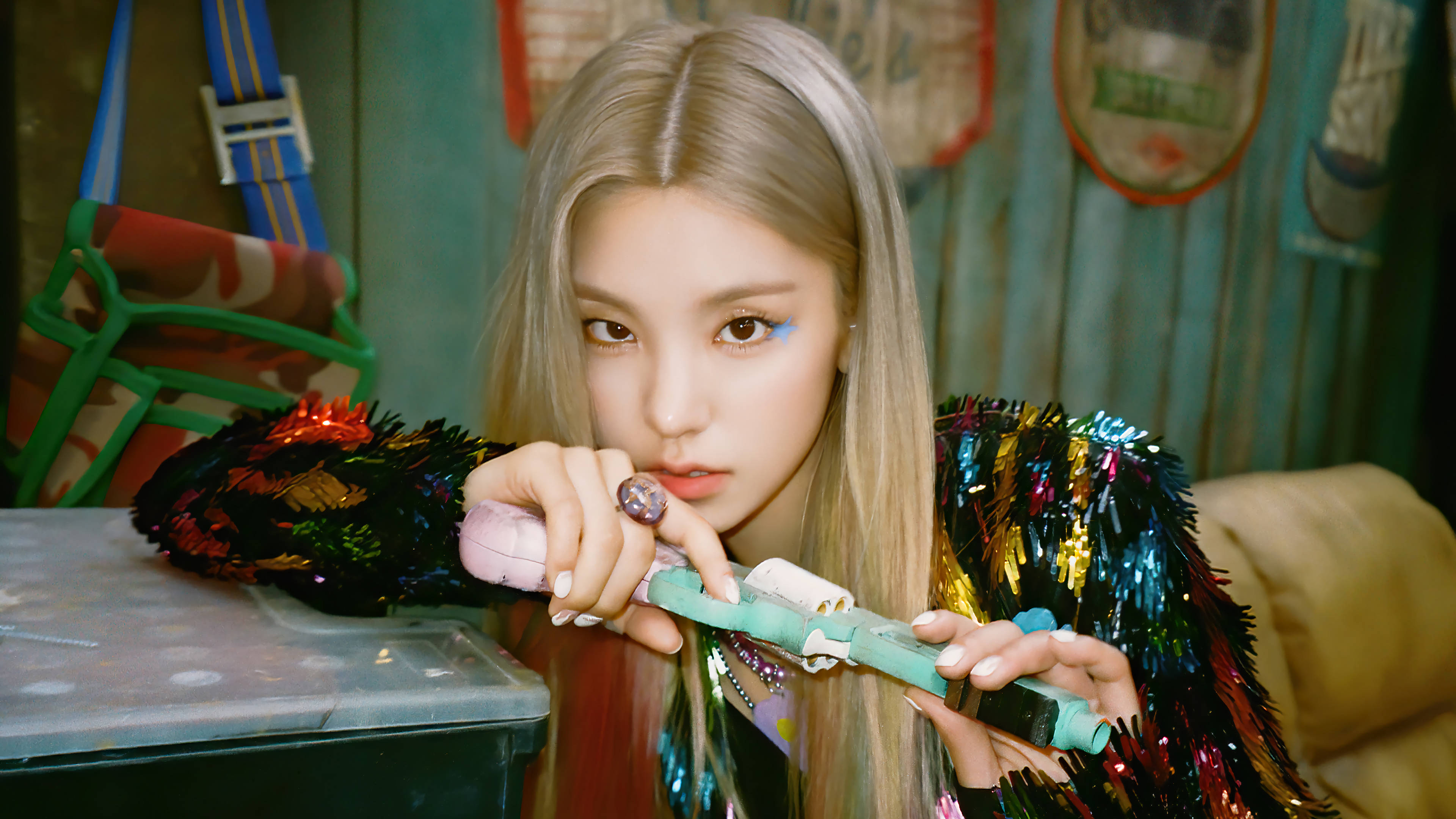 ITZY 桌布 壁紙 — 一位金髮碧眼的年輕女子，化著大膽的藍色眼妝，穿著閃亮的金屬絲夾克，手持彩色玩具槍。