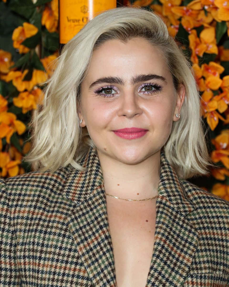 Mae Whitman 桌布 壁紙 — 一位金髮粉唇的女性在鮮豔的橙色花卉背景前溫暖地微笑。