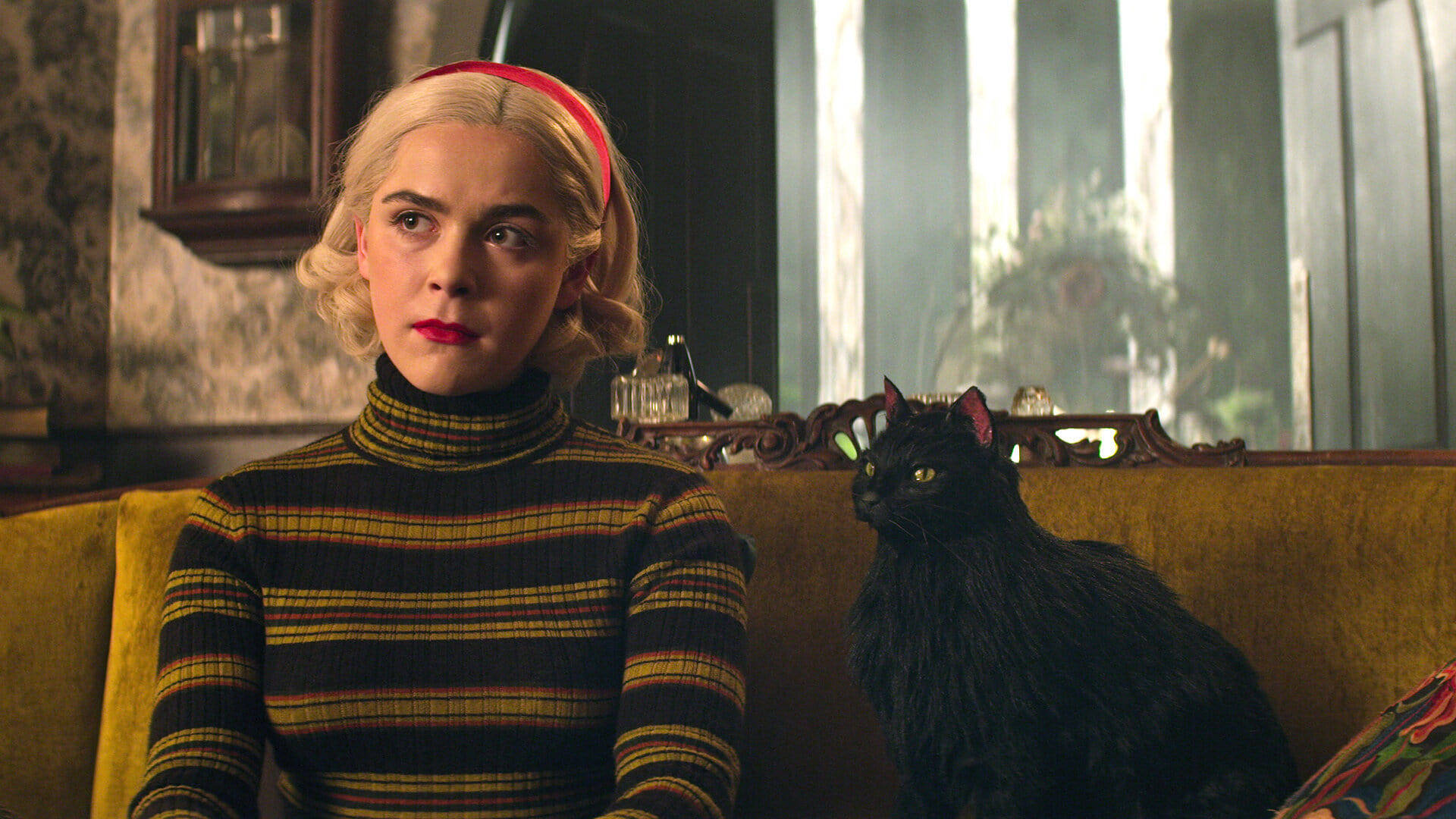 Kiernan Shipka 桌布 壁紙 — 一位金髮紅唇的女性坐在黃色沙發上，旁邊有一隻黑貓，身處裝飾華麗的復古房間。
