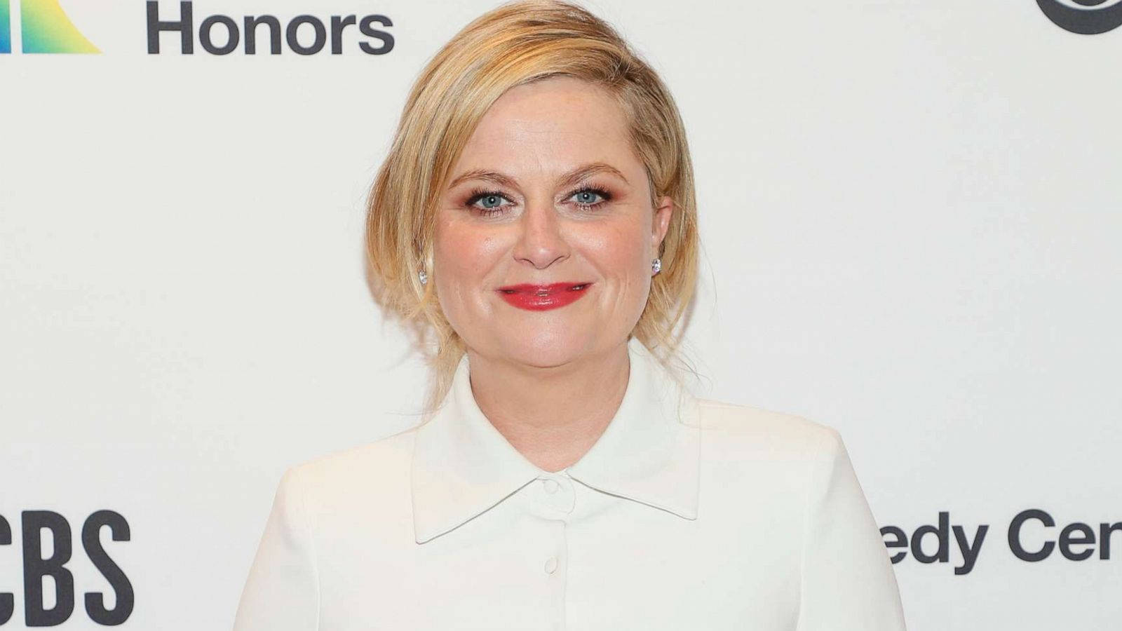oscars 桌布 壁紙 — 一位金髮紅唇的女性穿著白色有領襯衫，在帶有CBS和Comedy Central標誌的背景前微笑。