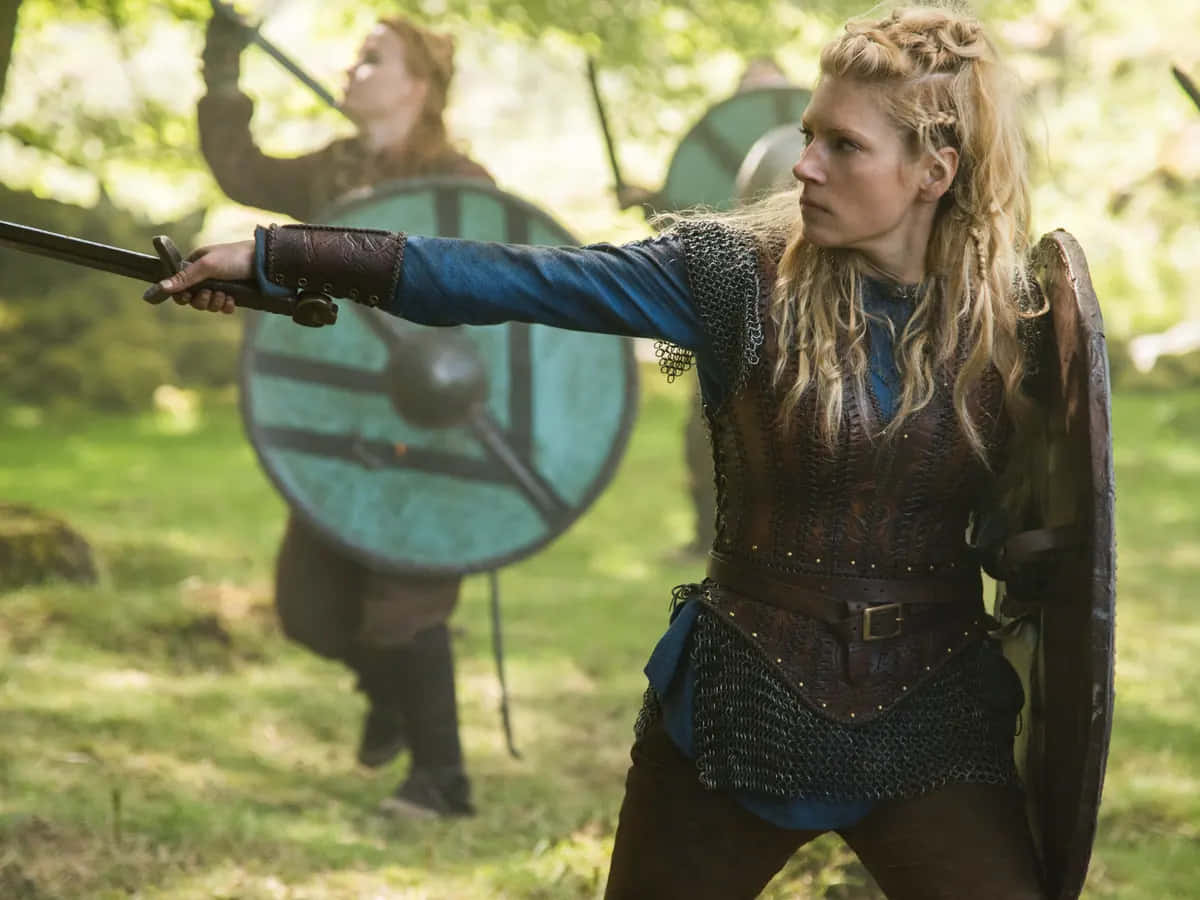 Female Viking Warriors 桌布 壁紙 — 一位金髮維京女子身穿鎖子甲和皮革盔甲，在陽光明媚的森林空地中準備好劍和盾牌。