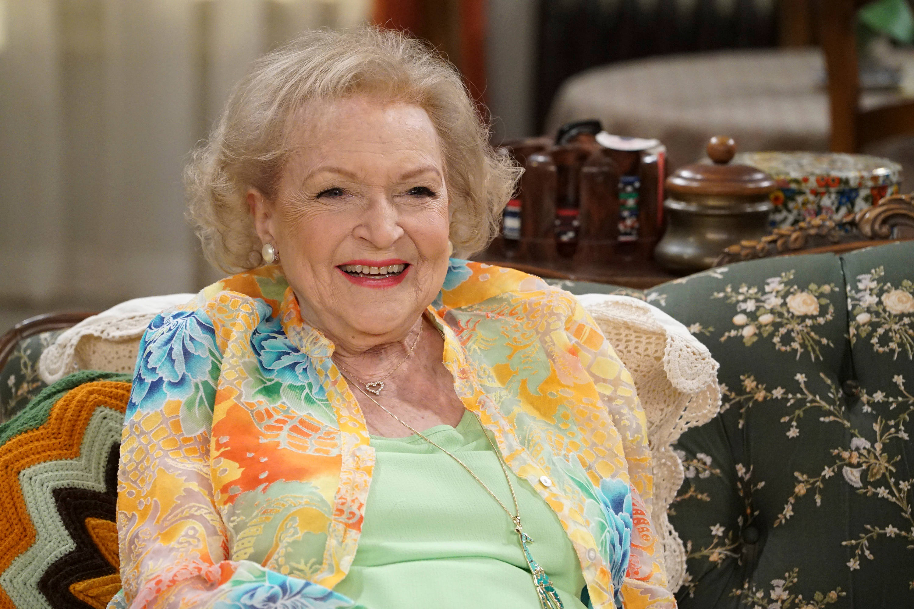 Betty White 桌布 壁紙 — 一位金髮老婦人坐在花卉圖案的沙發上，穿著色彩繽紛的花卉披肩，溫暖地微笑。