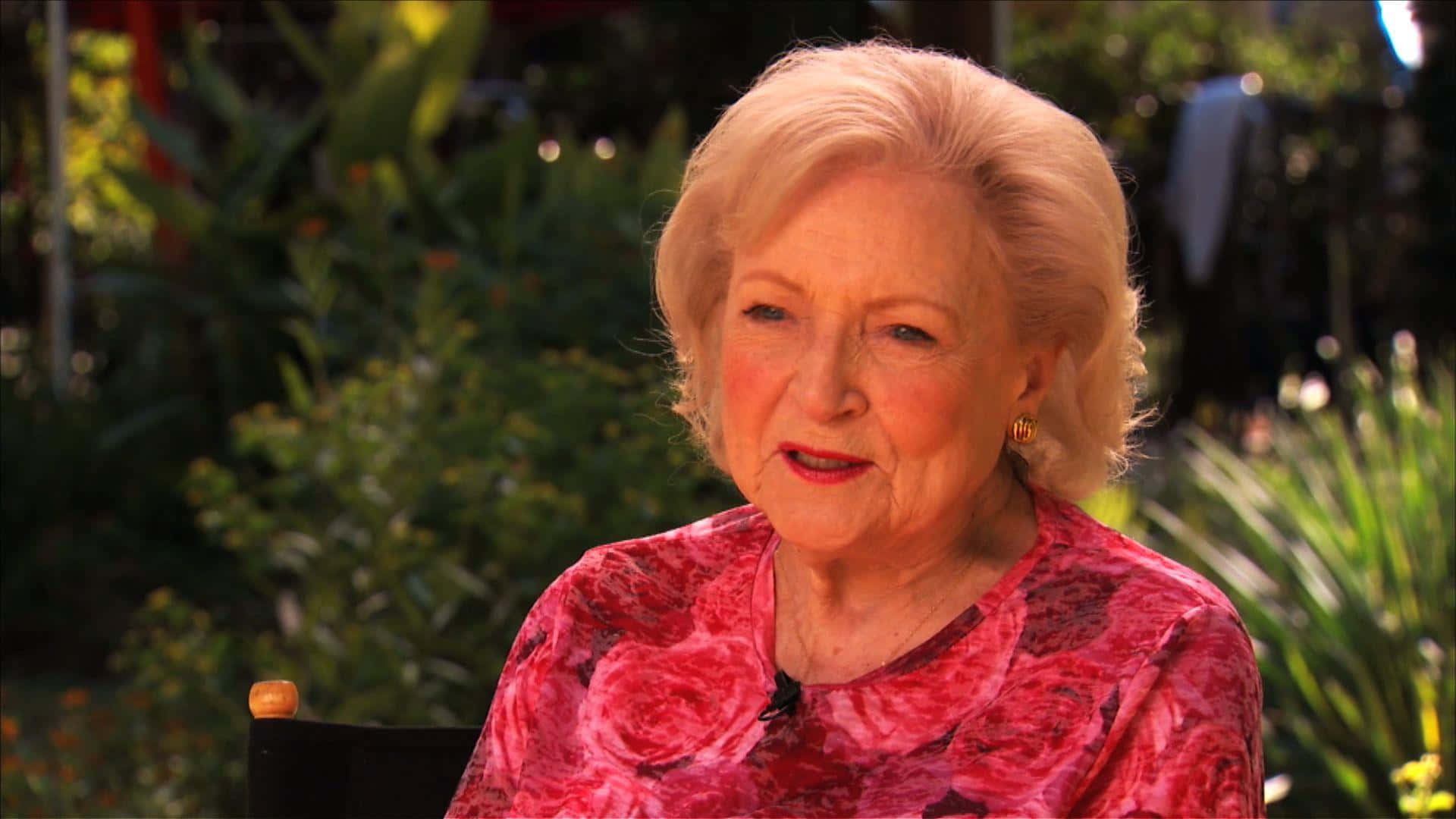 Betty White 桌布 壁紙 — 一位金髮老婦人穿著紅色花卉圖案襯衫，坐在戶外，周圍環繞著茂密的綠色植物。