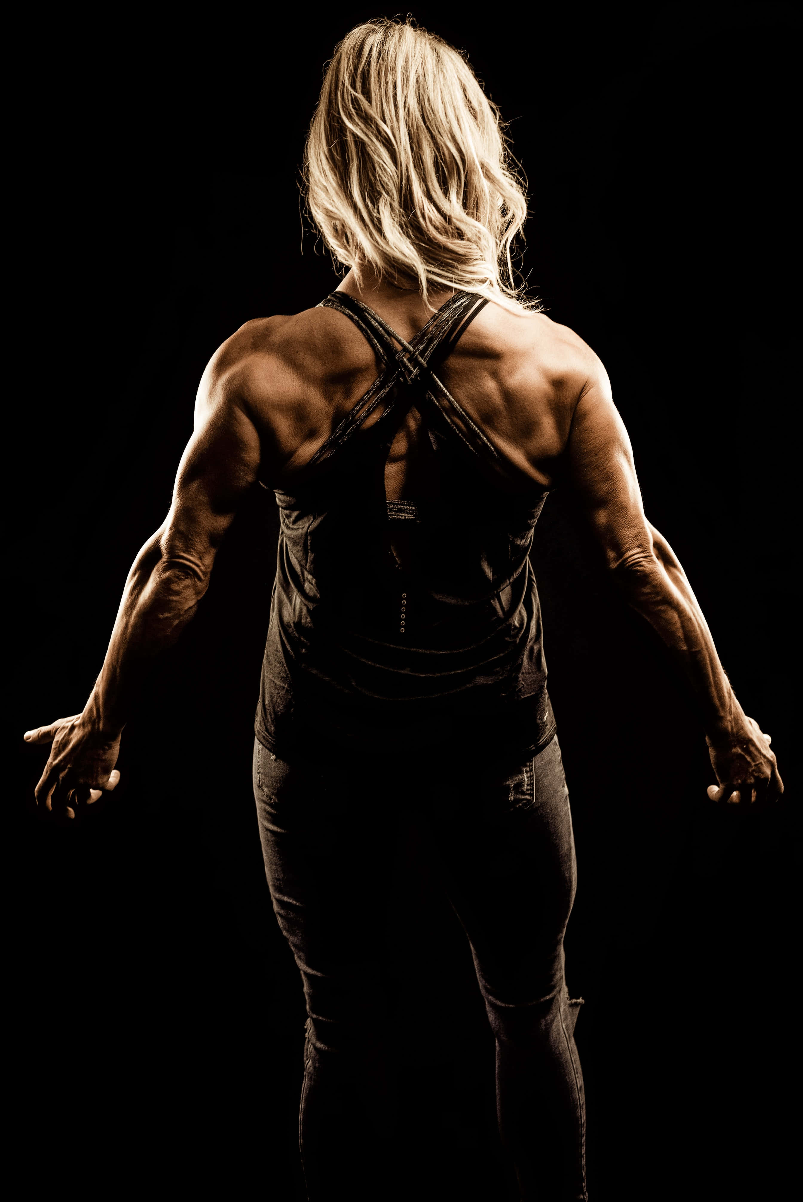 muscle growth 桌布 壁紙 — 一位金髮肌肉女性背光站在黑色背景前，在戲劇性的工作室拍攝中展示清晰的背部和手臂肌肉。