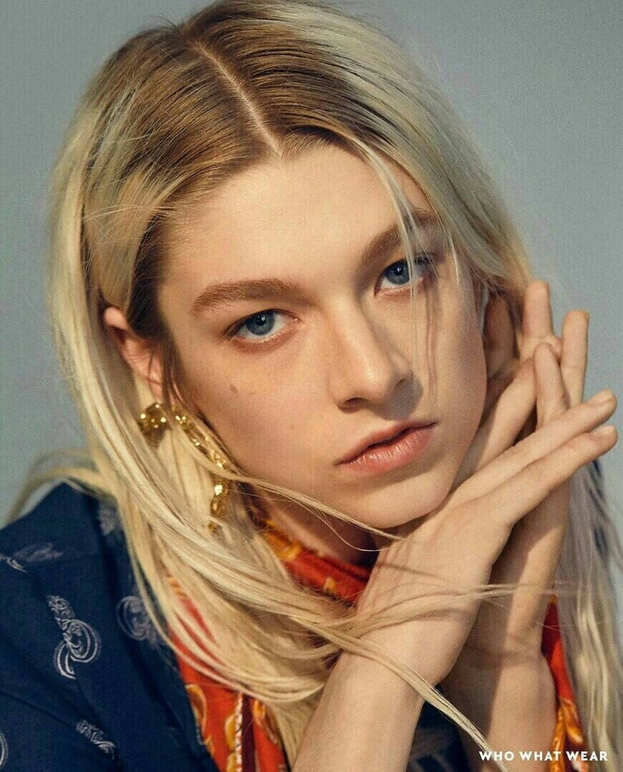 Hunter Schafer 桌布 壁紙 — 一位金髮藍眼女士雙手靠近臉龐，在柔和灰色背景前直視鏡頭。