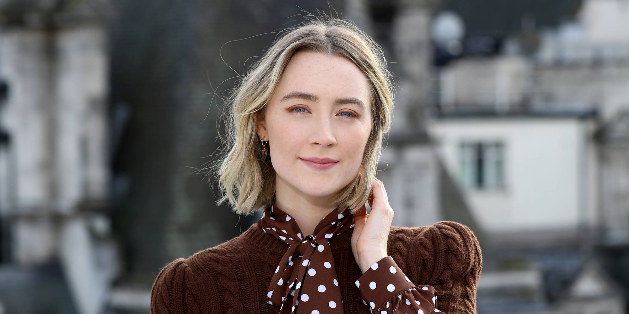 Saoirse Ronan 桌布 壁紙 — 一位金髮藍眼的女人輕柔地微笑，同時觸摸她的脖子，穿著棕色麻花針織毛衣和波點圍巾，背景是模糊的城市景觀。